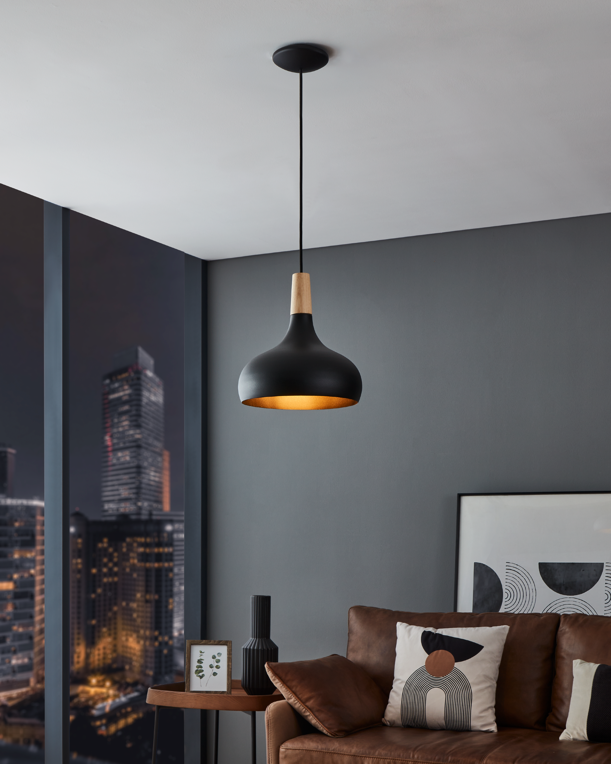 Una lampada a sospensione nera è appesa in una stanza. Un divano in pelle marrone, un tavolo con un quadro e vista sulla città sono visibili.