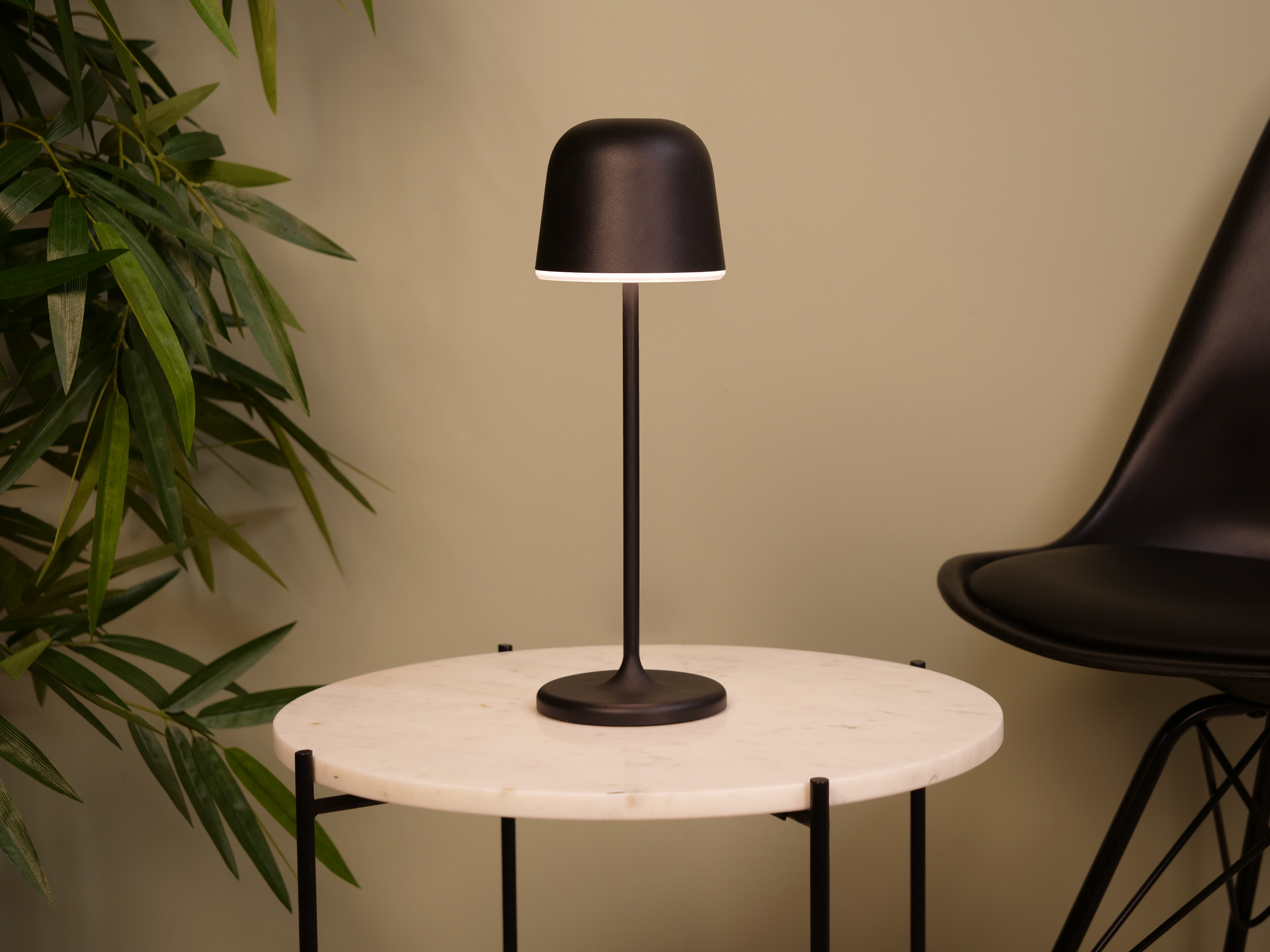 Eine schwarze Lampe auf einem Marmortisch, Bambusblätter links und ein schwarzer Stuhl rechts.