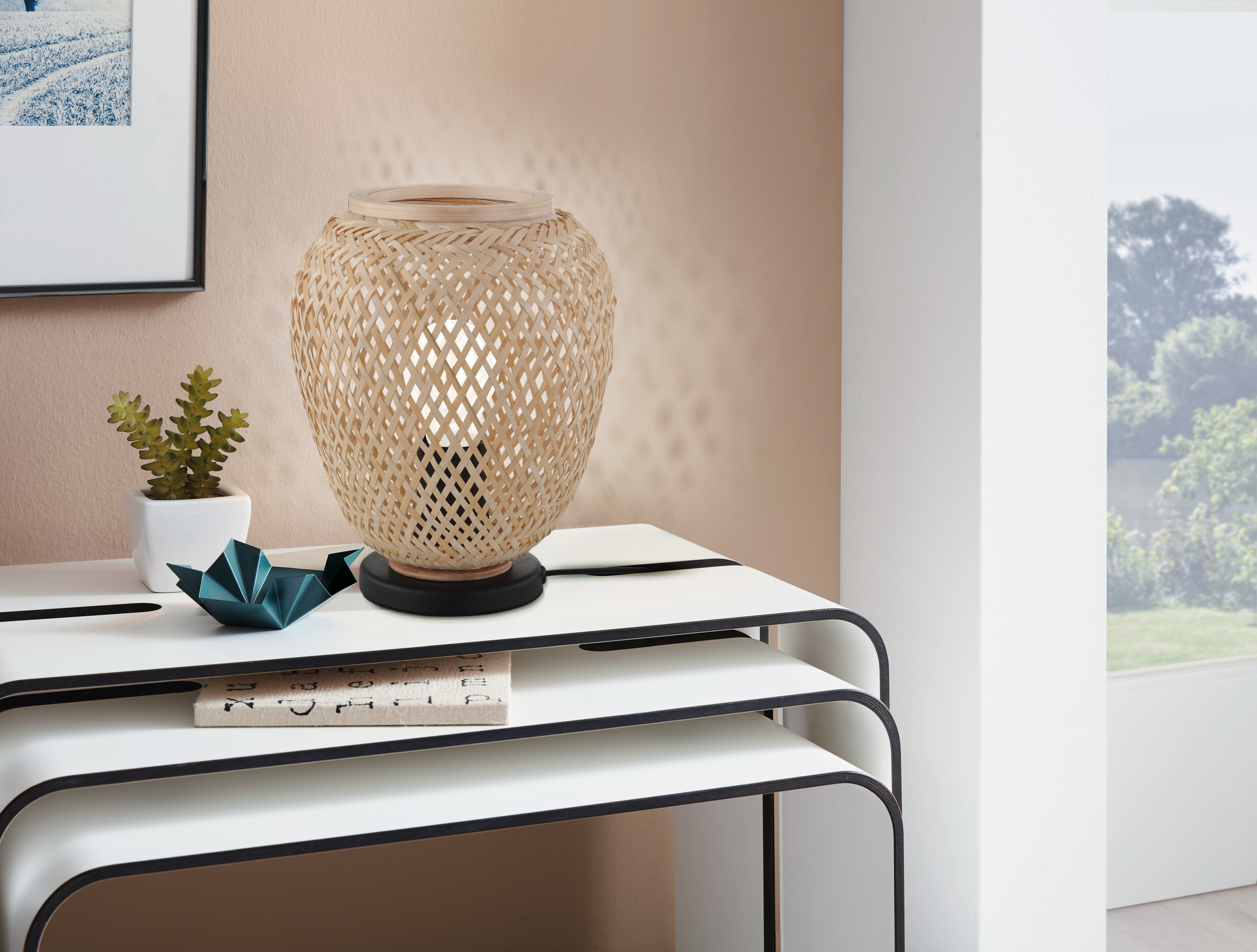 Eine gewebte Lampe auf einem Tisch mit einer Pflanze und Origami. Der Hintergrund hat ein Fenster mit Bäumen und einer Wand.