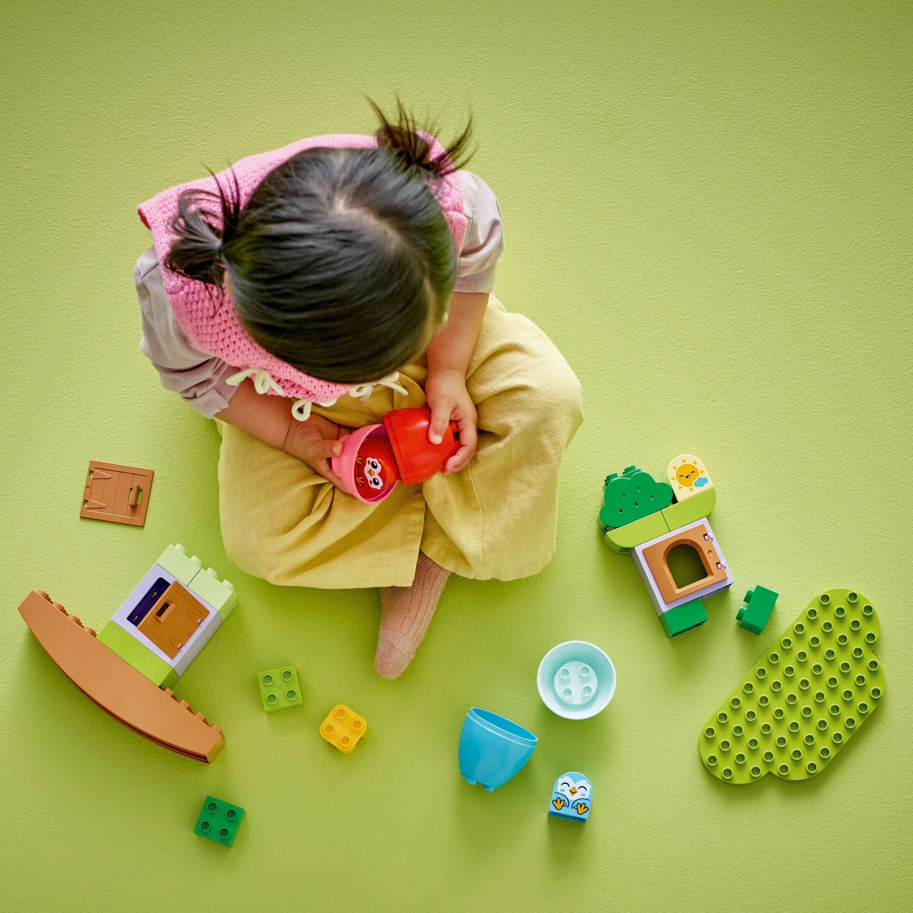 Dziewczynka bawiąca się klockami Lego Duplo. Klocki zawierają domek na drzewie, niebieską sowę i inne kolorowe elementy.