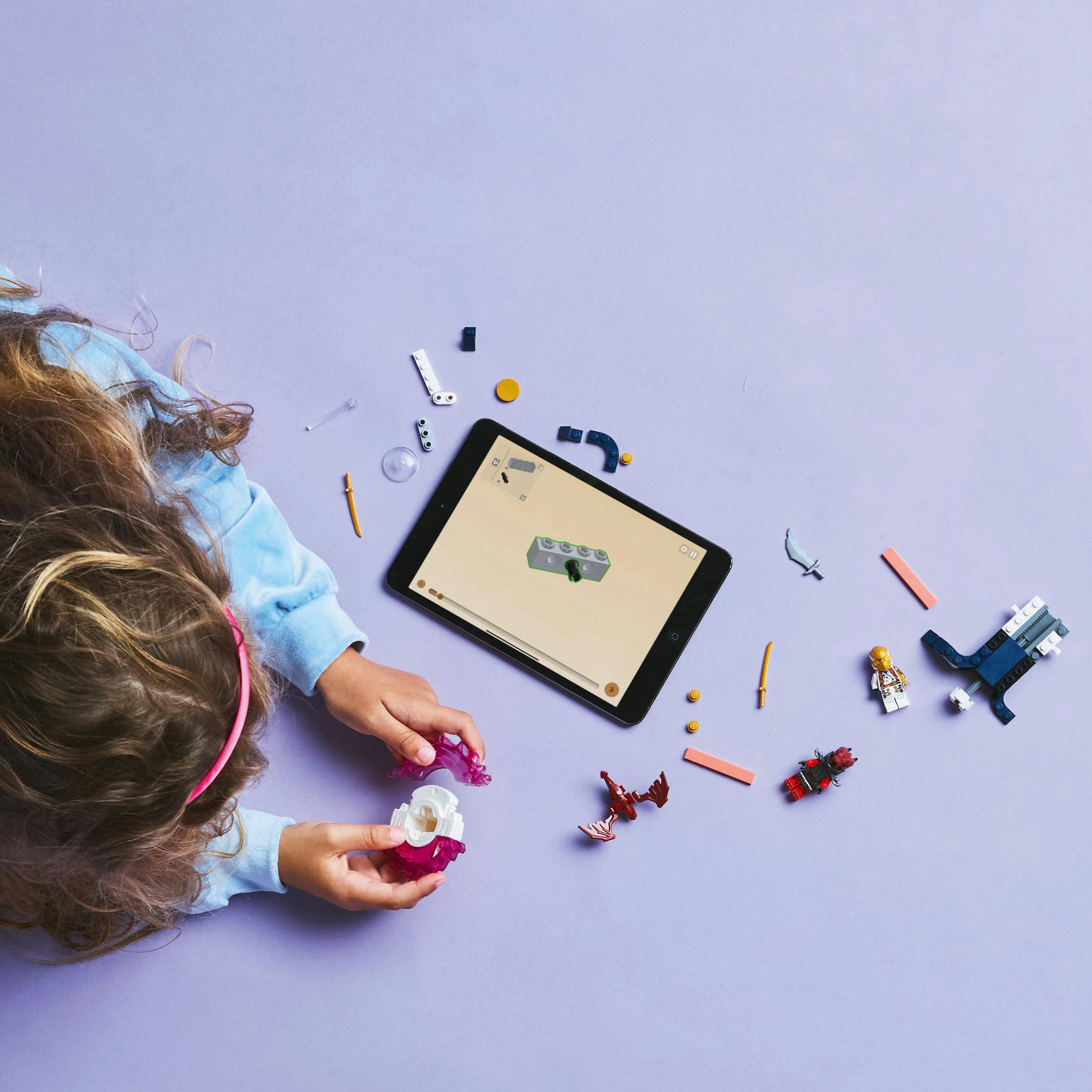 Handen die Lego-stenen monteren op een paars oppervlak. Een tablet met instructies en andere Lego-onderdelen zijn zichtbaar.