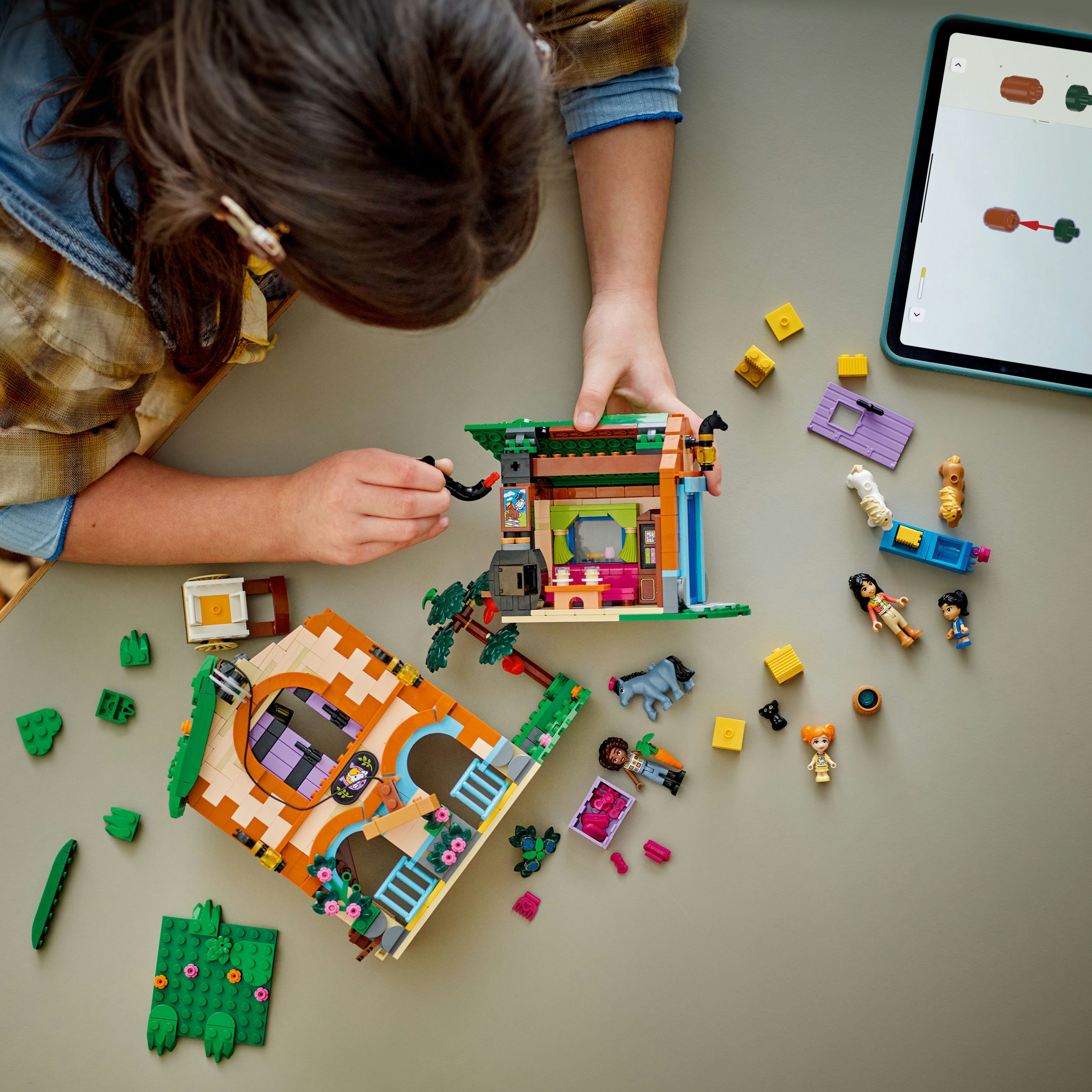 Bir kişi, hayvan figürleriyle bir Lego ev inşa ediyor ve gri bir yüzeyde talimatları gösteren bir tablet var.