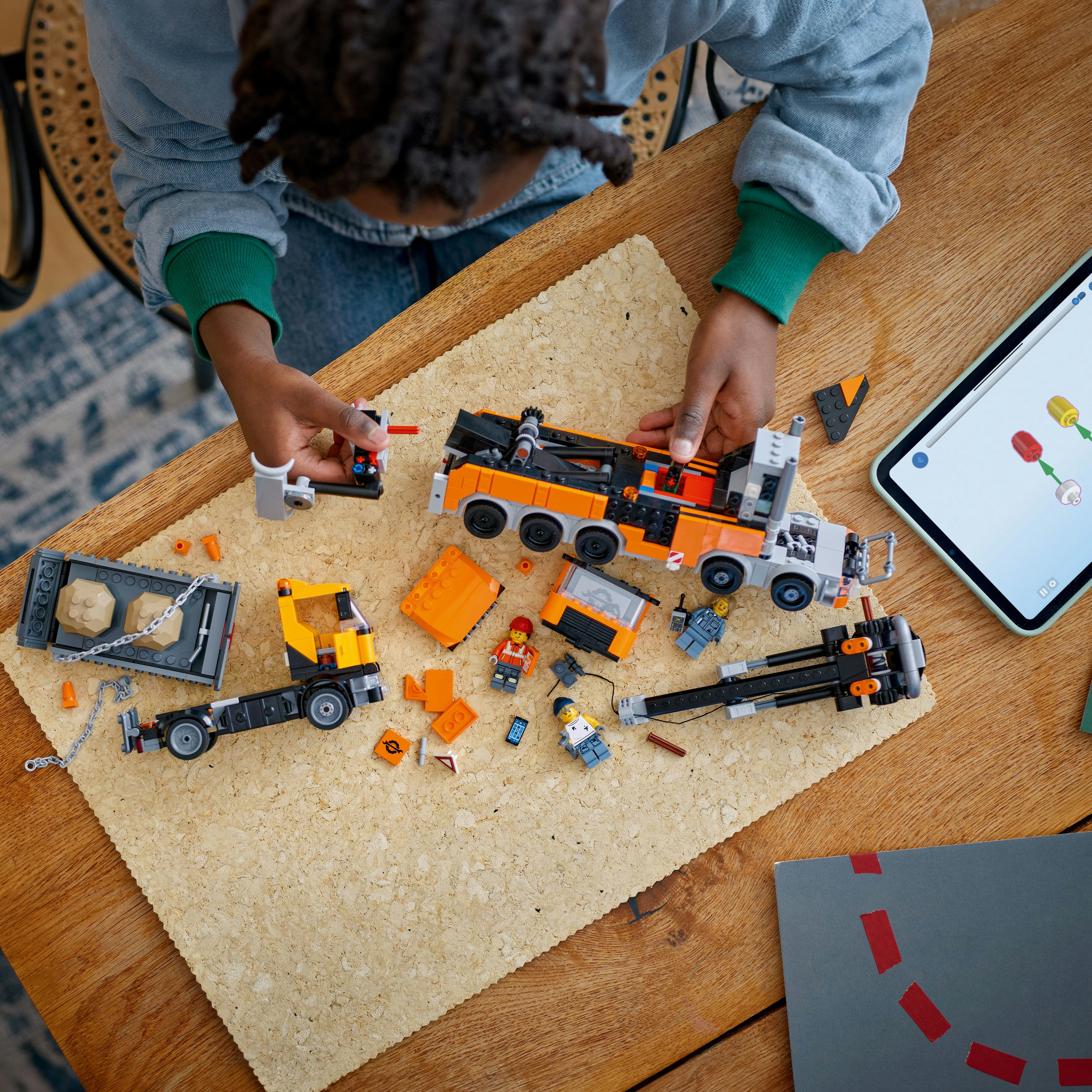 Dziecko składa lawetę LEGO, z częściami i tabletem. Pomarańczowe, szare i żółte elementy LEGO na brązowej powierzchni.