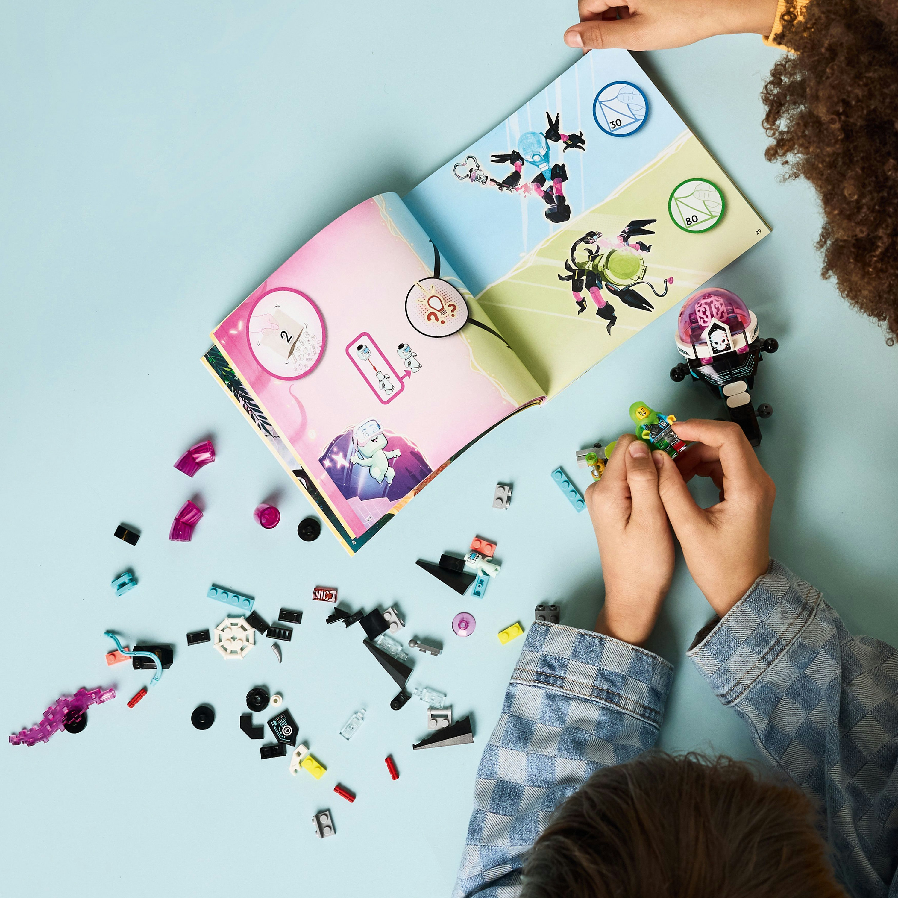 Dos niños juegan con piezas de Lego y un libro de Lego sobre una superficie azul.