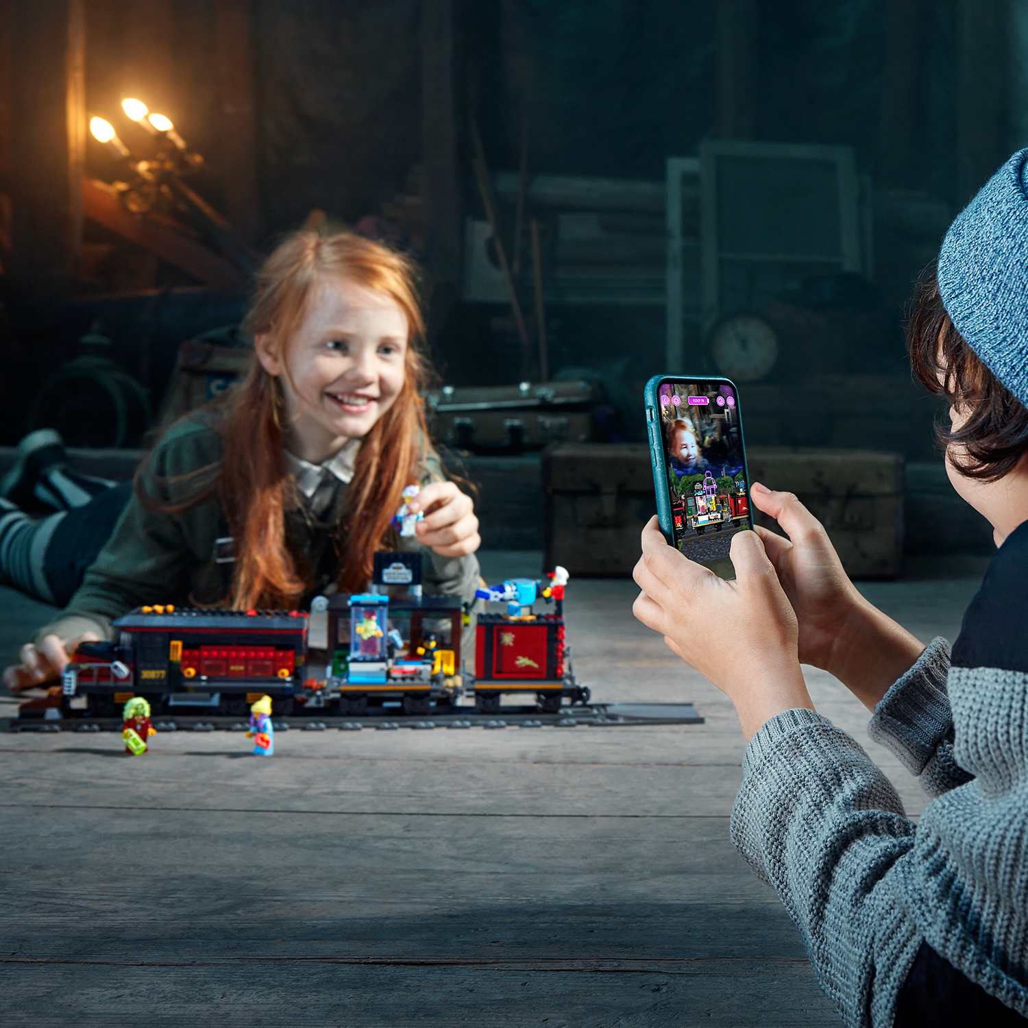 Dwoje dzieci bawiących się zestawem pociągu Lego, jedno robi zdjęcie telefonem.