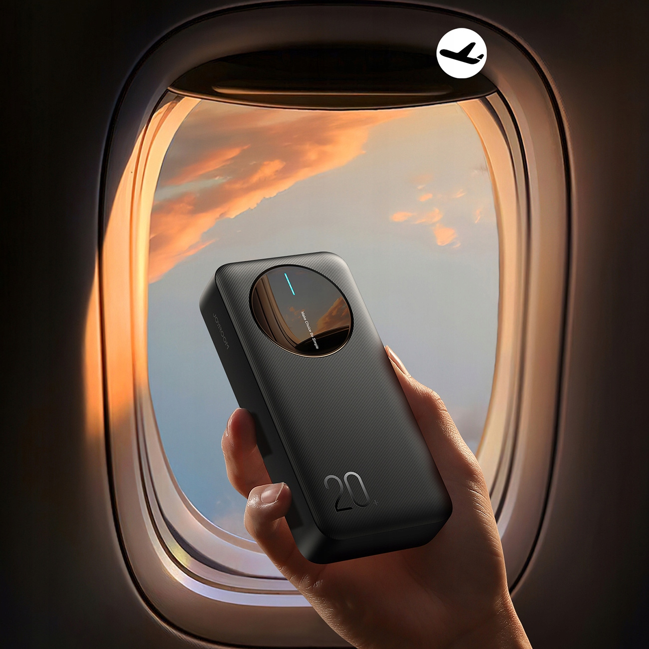 Czarny power bank trzymany jest w pobliżu okna samolotu, ukazując zachodzące słońce.