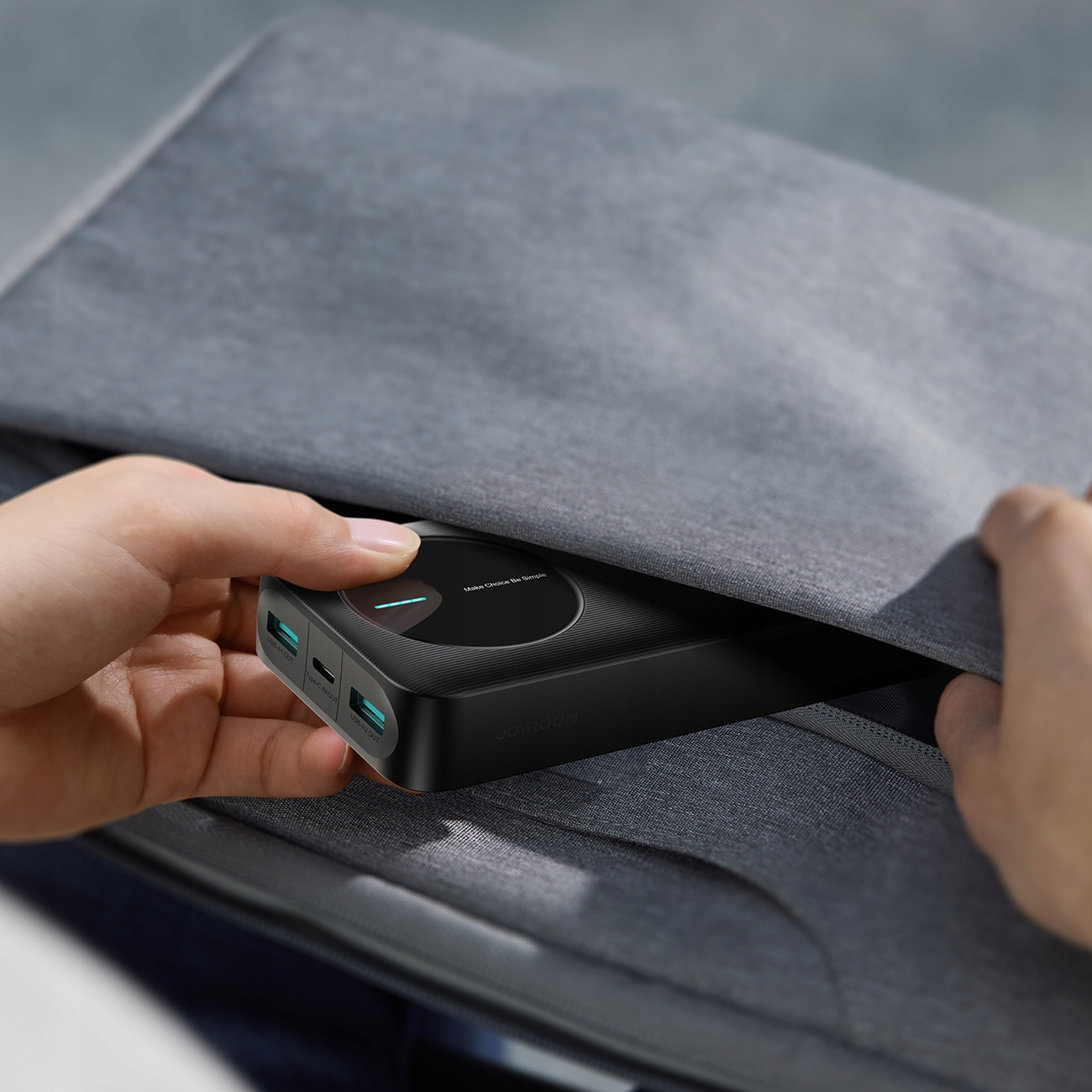 Ręce wkładają czarny power bank z niebieskimi portami do szarej torby.