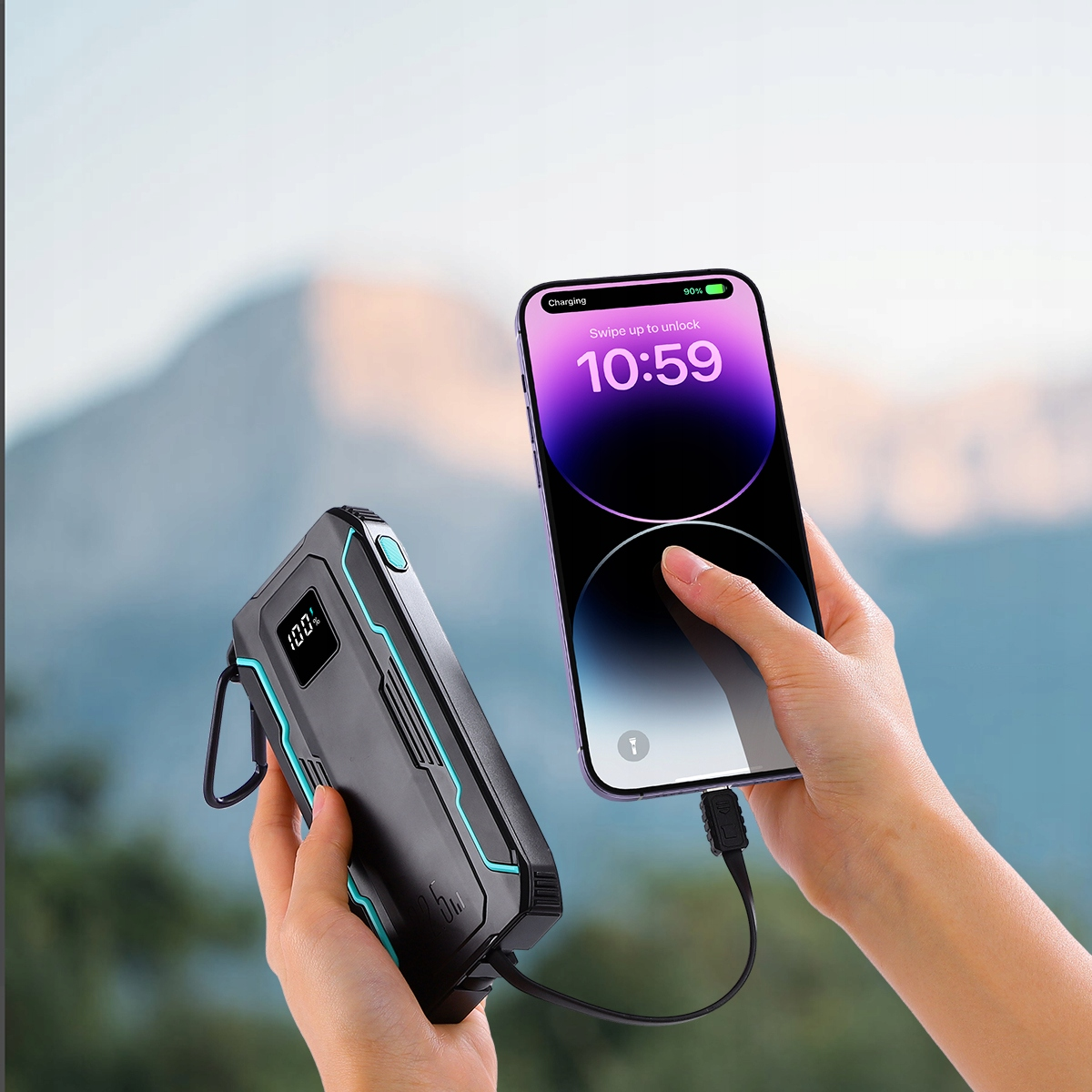 Eine Person hält eine schwarze Powerbank und ein Telefon, das über ein Kabel damit verbunden ist. Berge sind im Hintergrund.