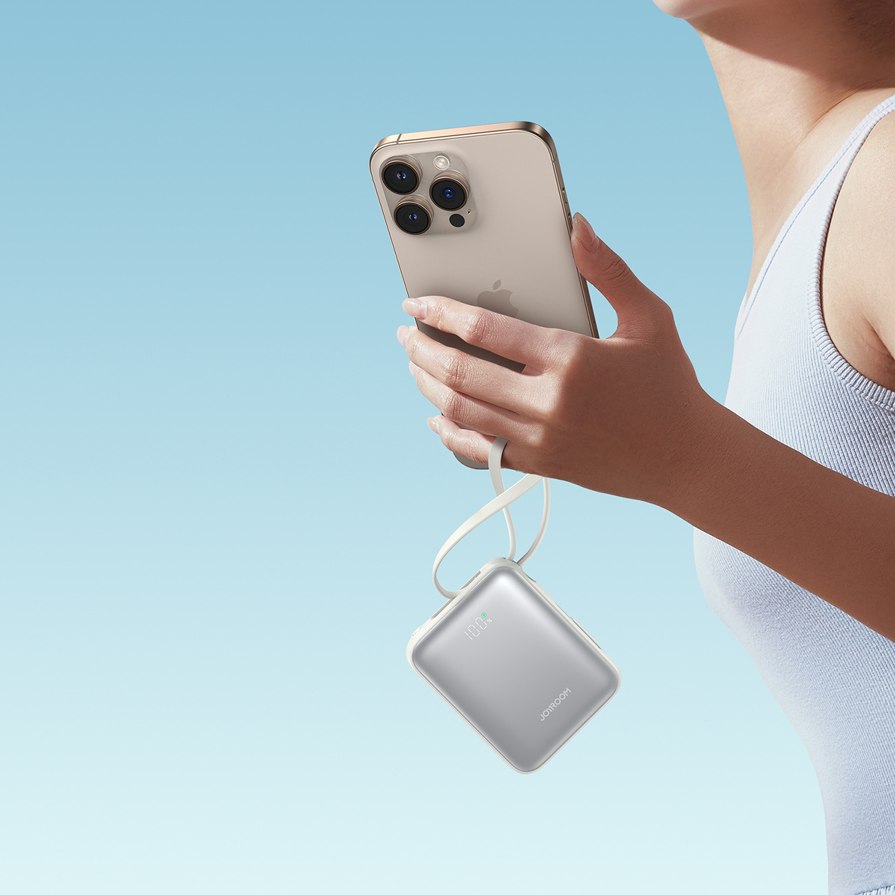 Kobieta trzyma telefon i srebrny power bank. Telefon się ładuje. Tło to błękitne niebo.