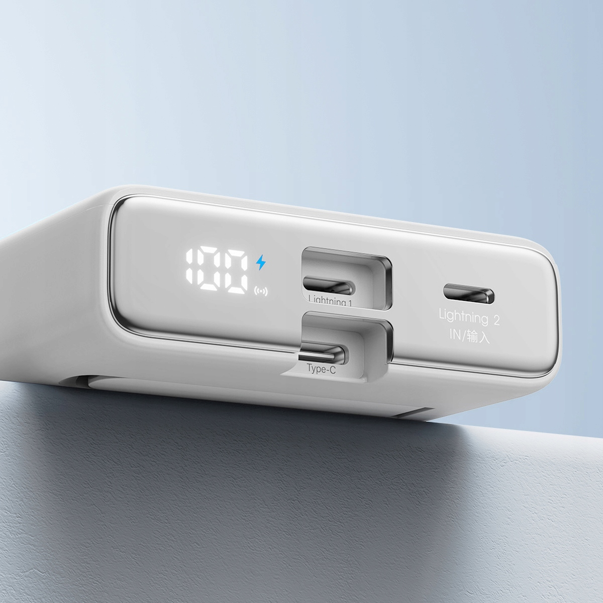 Biały power bank z wyświetlaczem pokazującym 100% oraz portami Lightning i Type-C. Znajduje się na szarej powierzchni.