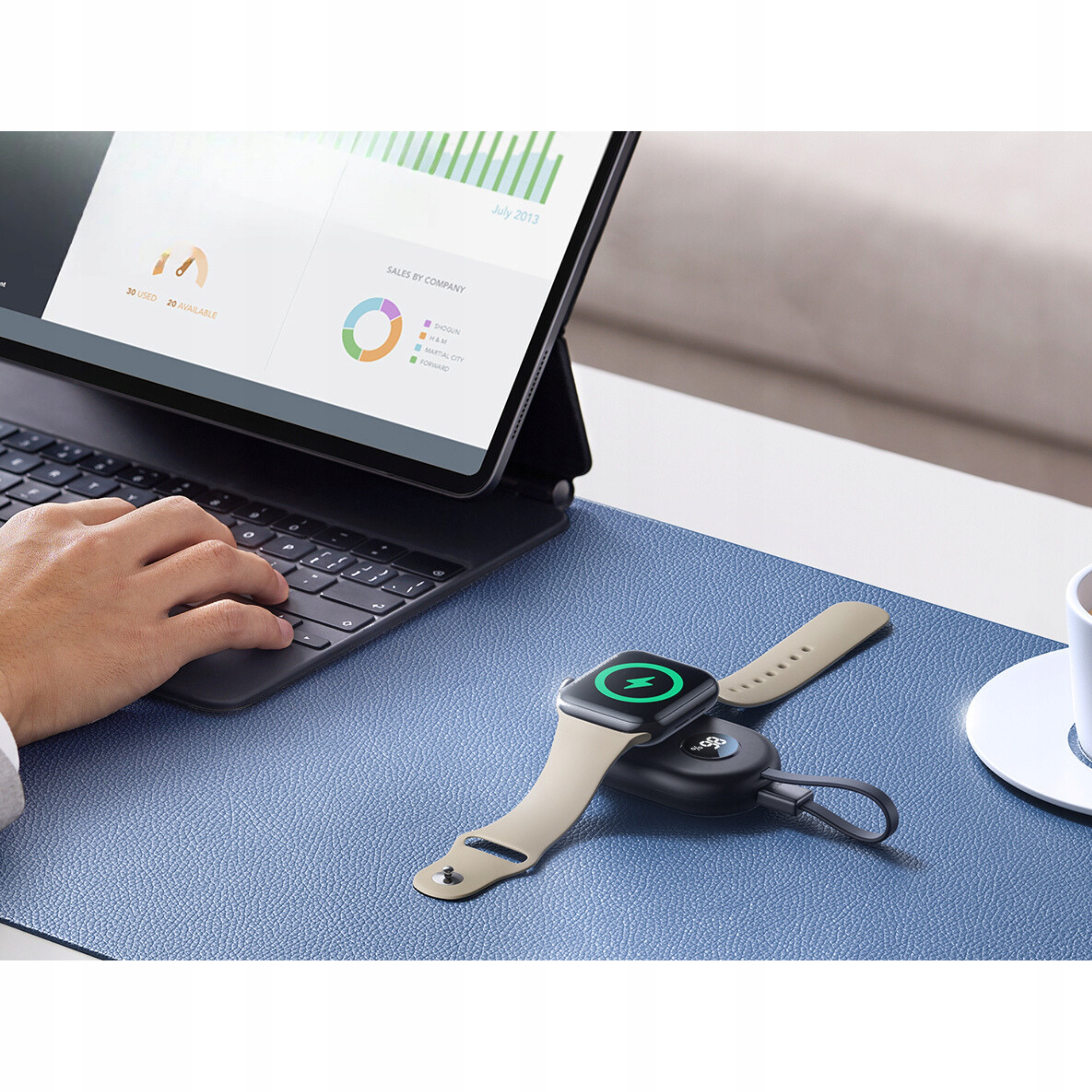 Osoba pisząca na klawiaturze, podkładka do ładowania bezprzewodowego ze smartwatchem na niebieskiej macie.