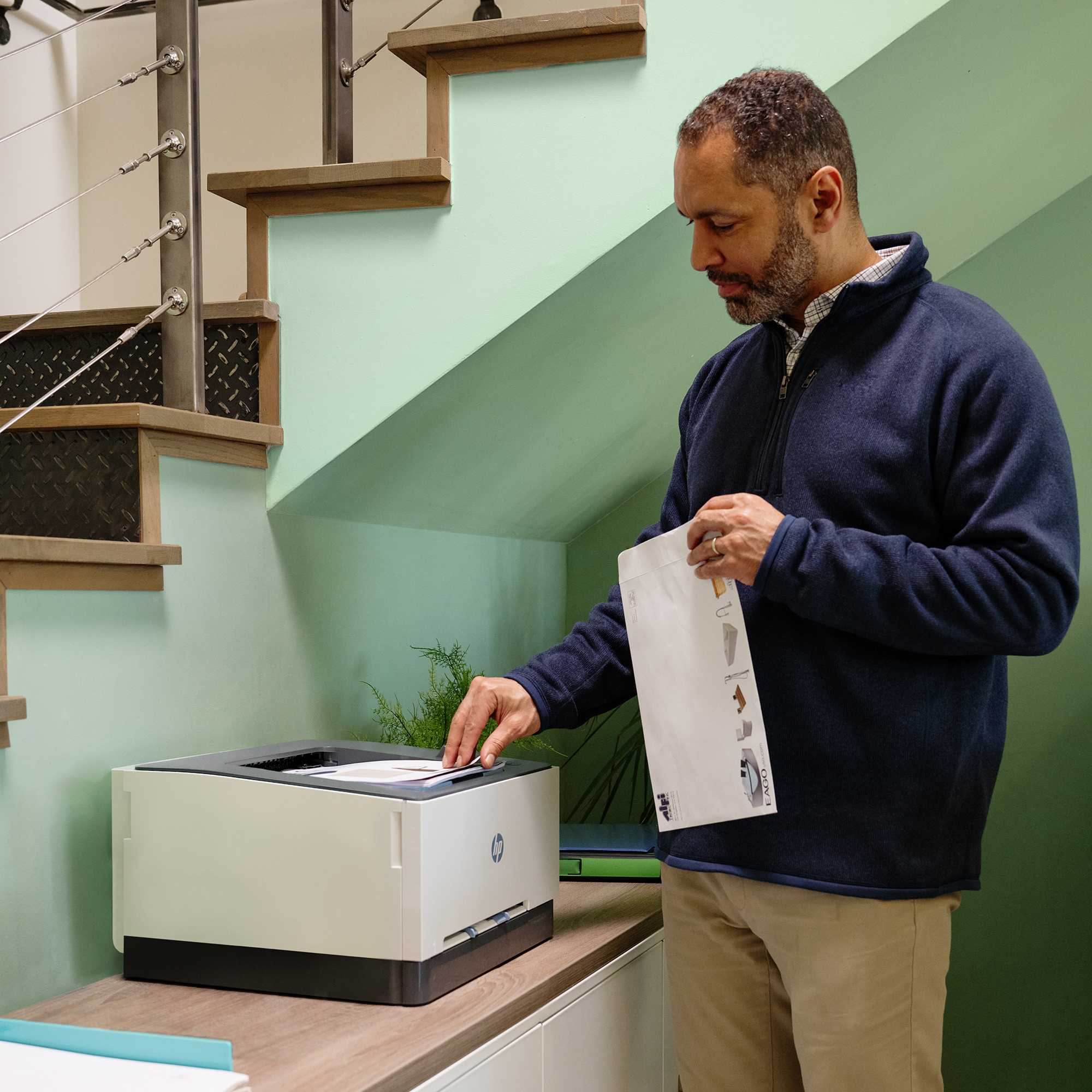 Un hombre con una chaqueta azul está cerca de una impresora, sosteniendo un documento. Se ve una pared verde y escaleras.