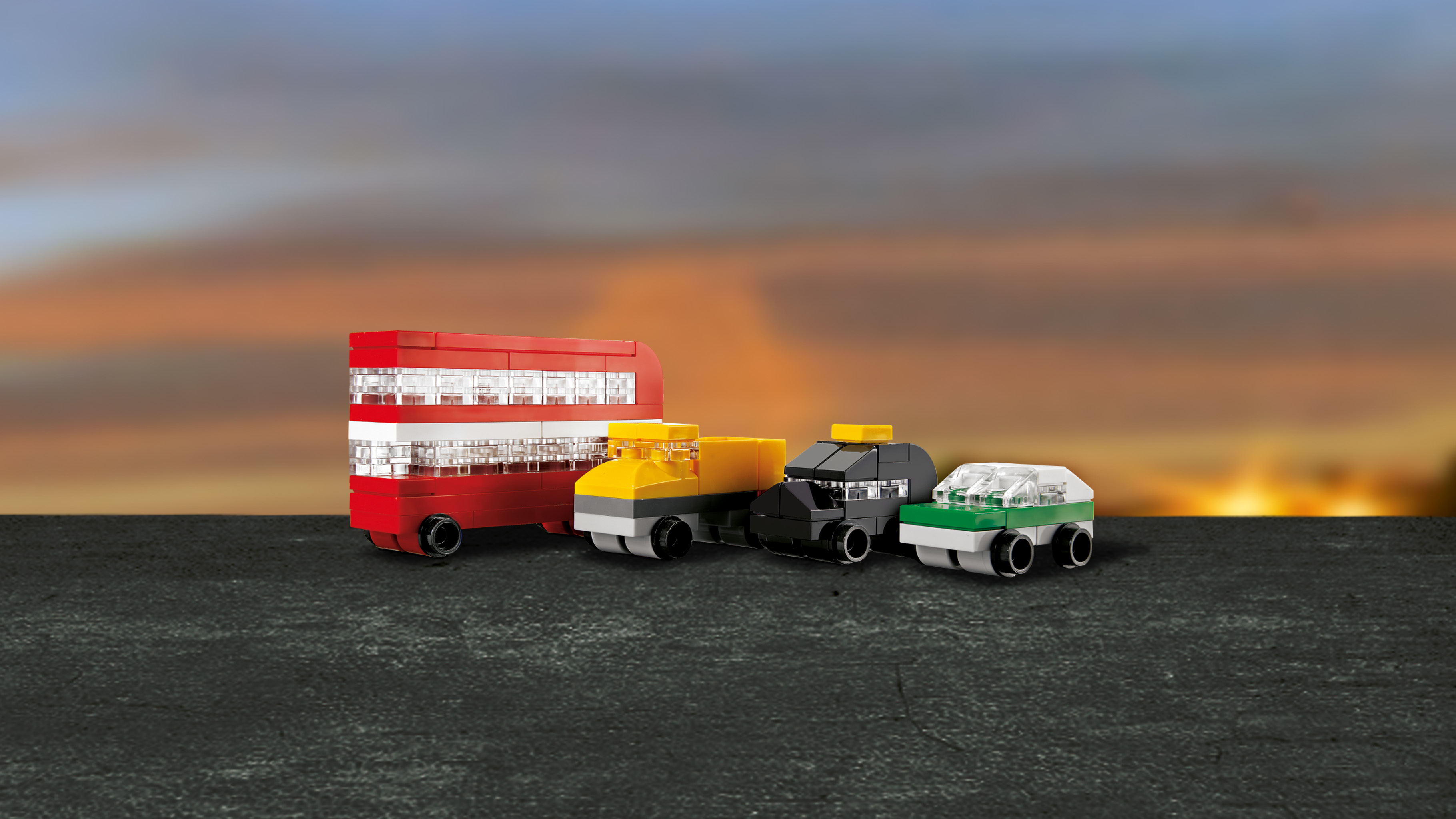 Cztery zabawki samochodowe na stole, w tym czerwony autobus piętrowy, żółta ciężarówka, czarny samochód i zielony samochód.