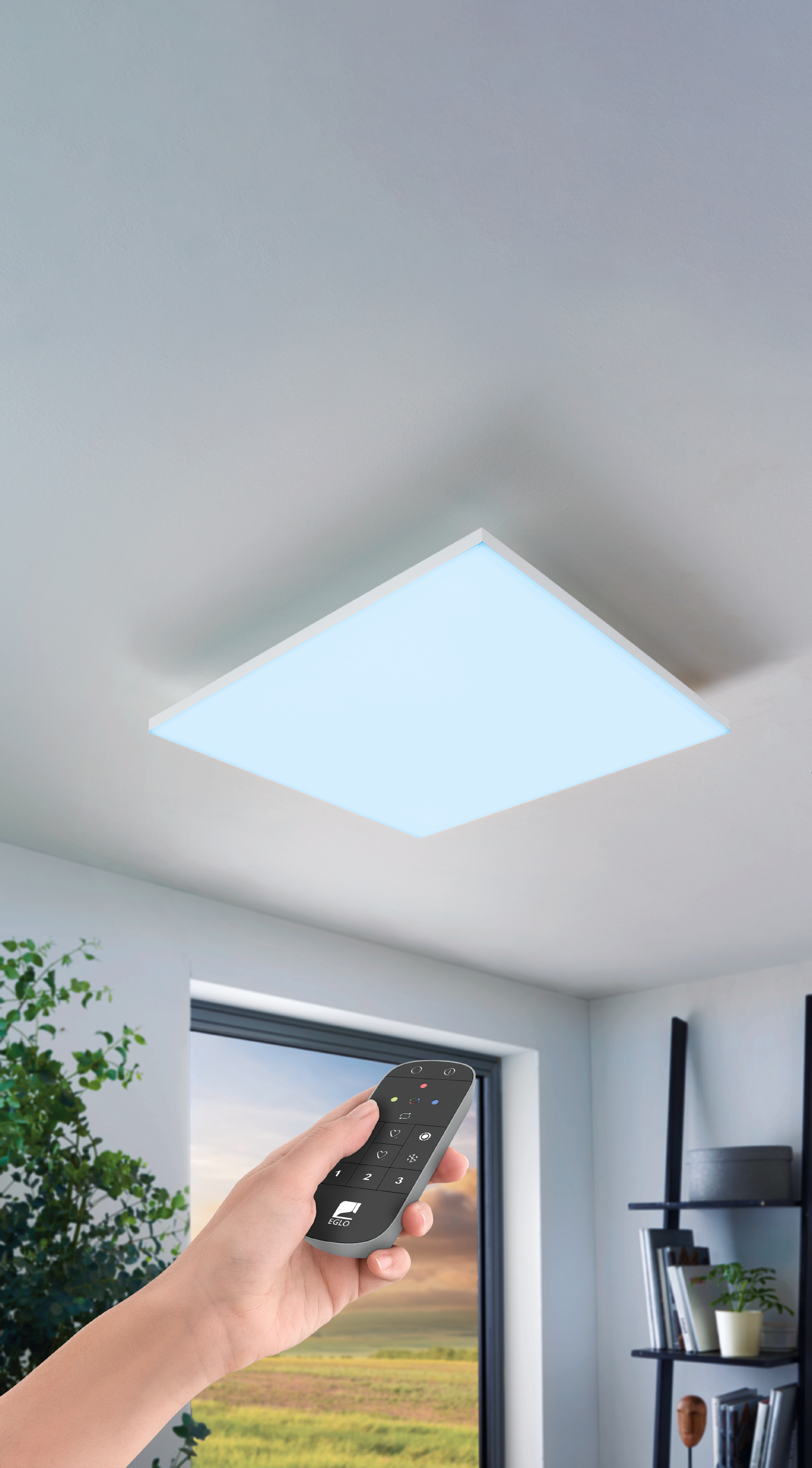 Una mano tiene un telecomando, puntando a una luce LED quadrata a soffitto in una stanza con finestra e scaffale.