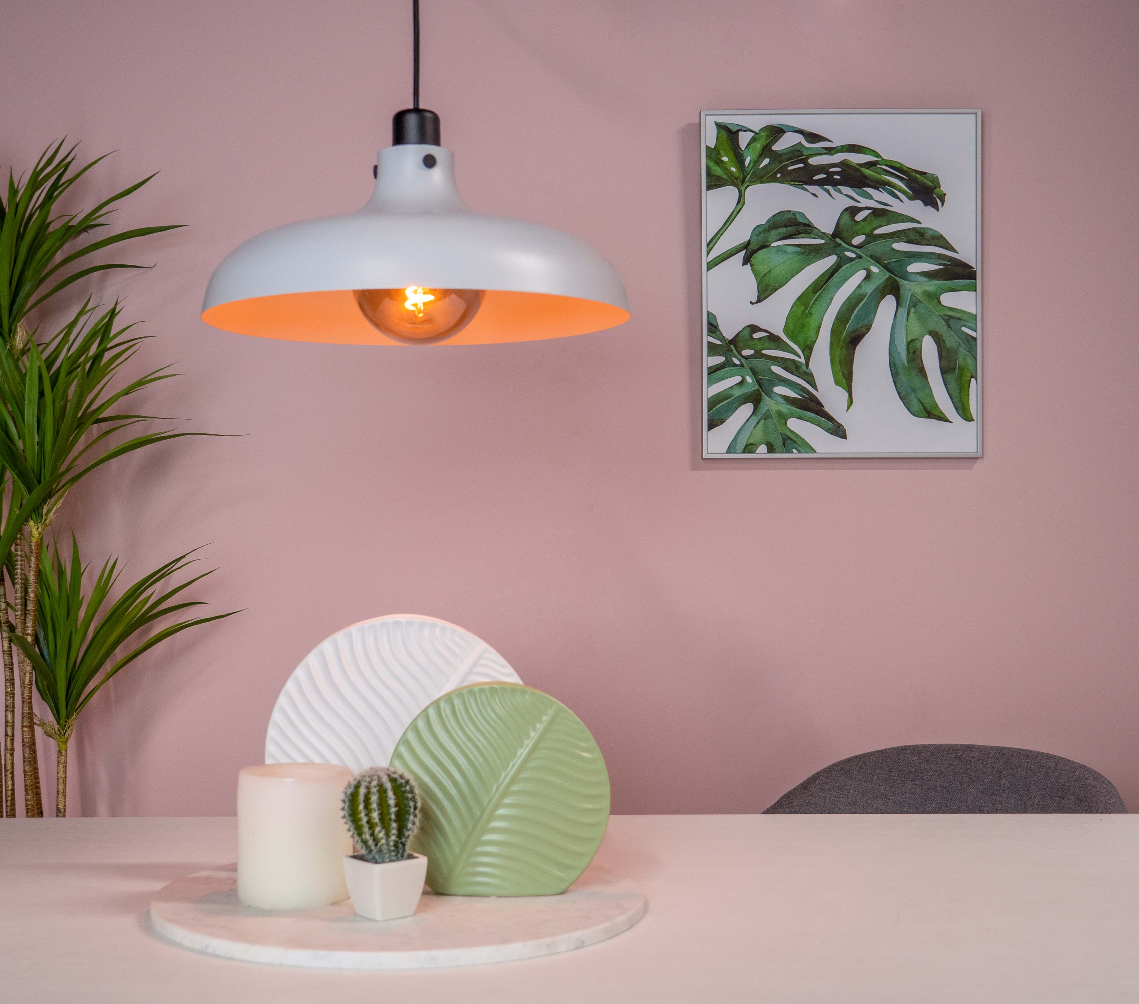 Weiße Hängelampe über einem Tisch mit Deko, ein Gemälde an rosa Wand. Warmes Licht.