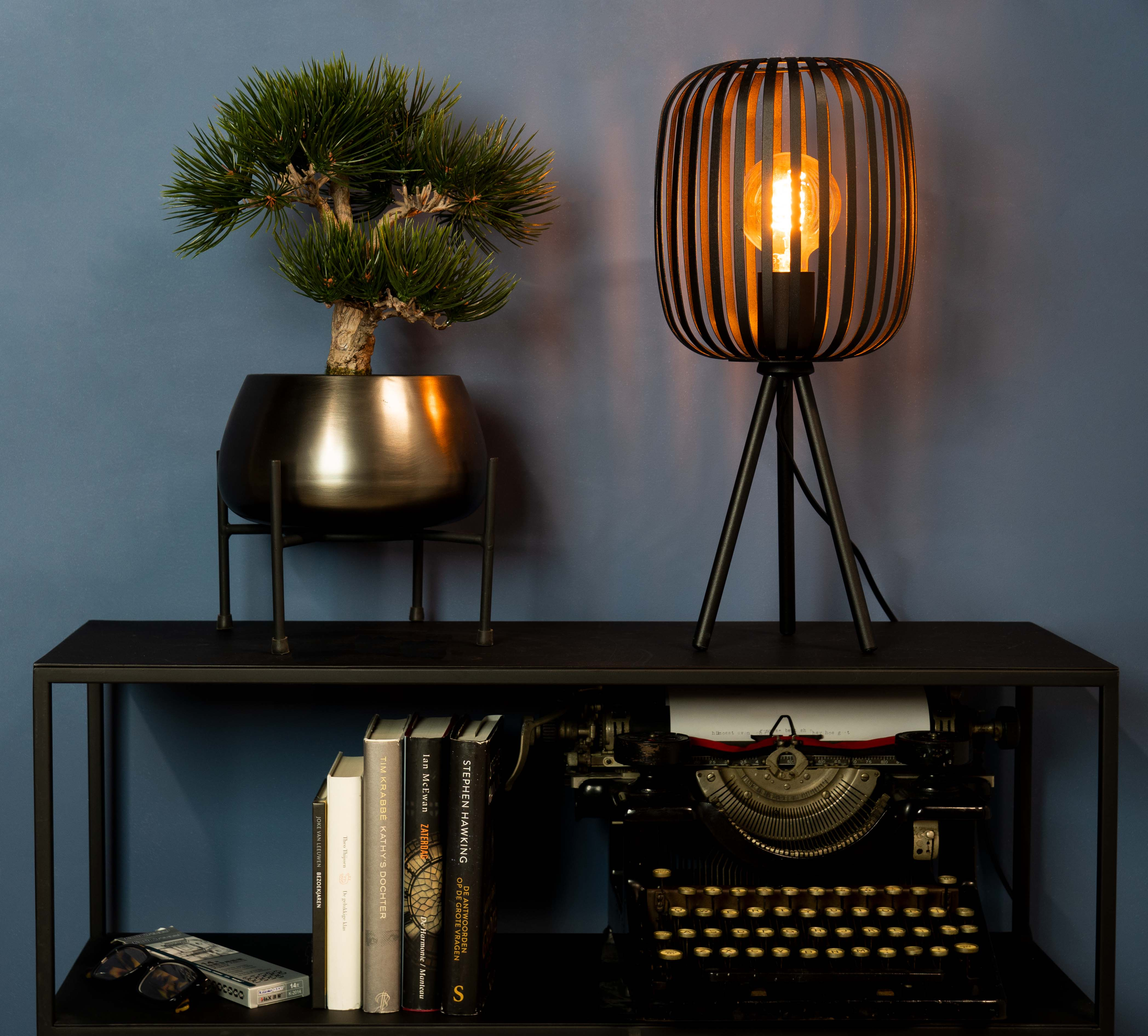Een zwarte tafel met een lamp, plant, boeken en typemachine tegen een blauwe muur.