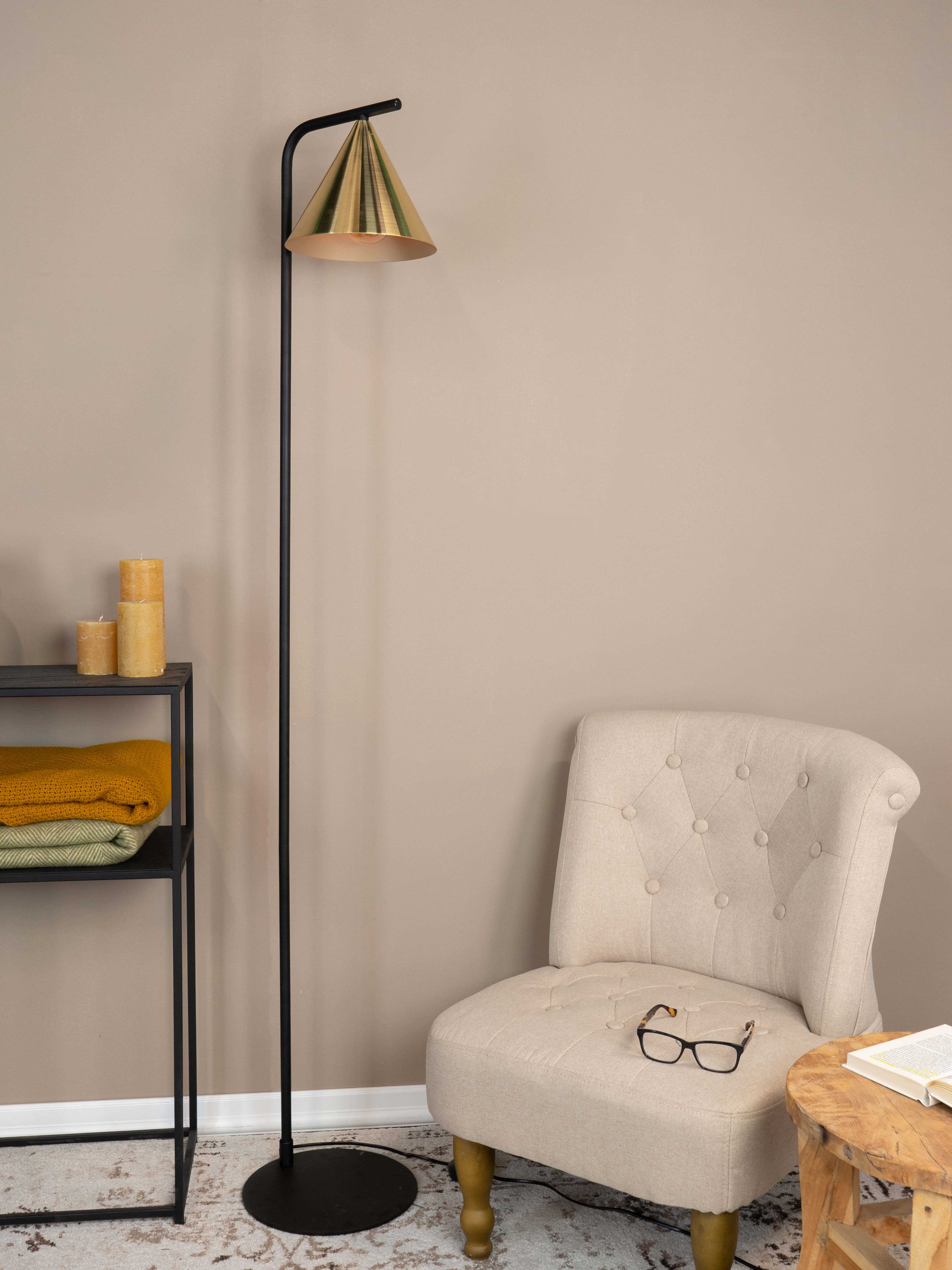 Eine Stehlampe mit goldenem Lampenschirm neben einem Stuhl, Regal und kleinem Tisch in einer inszenierten Raumumgebung.