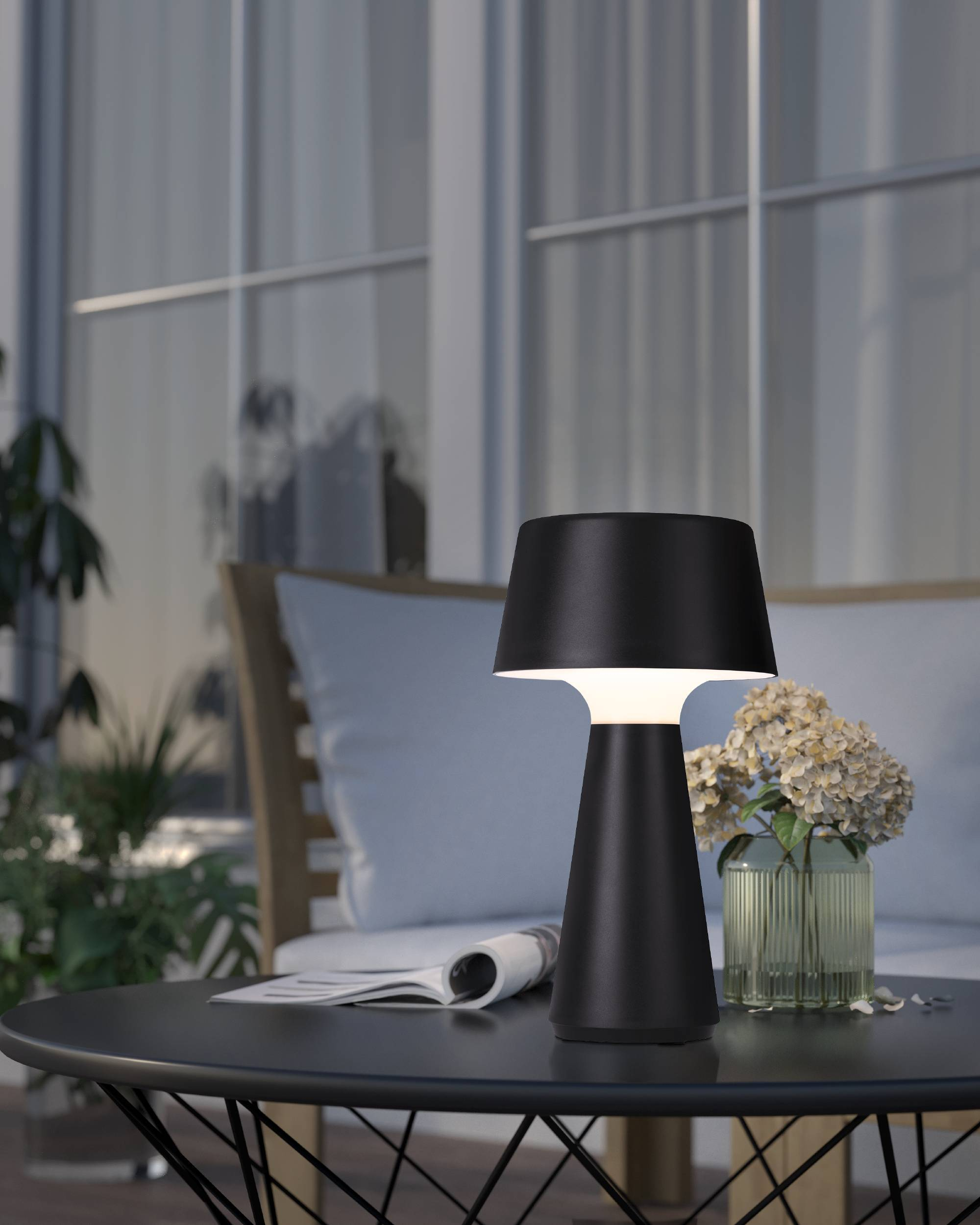 Eine schwarze Tischlampe auf einem schwarzen Tisch, im Freien auf einer Terrasse mit Pflanzen, Blumen und Möbeln.
