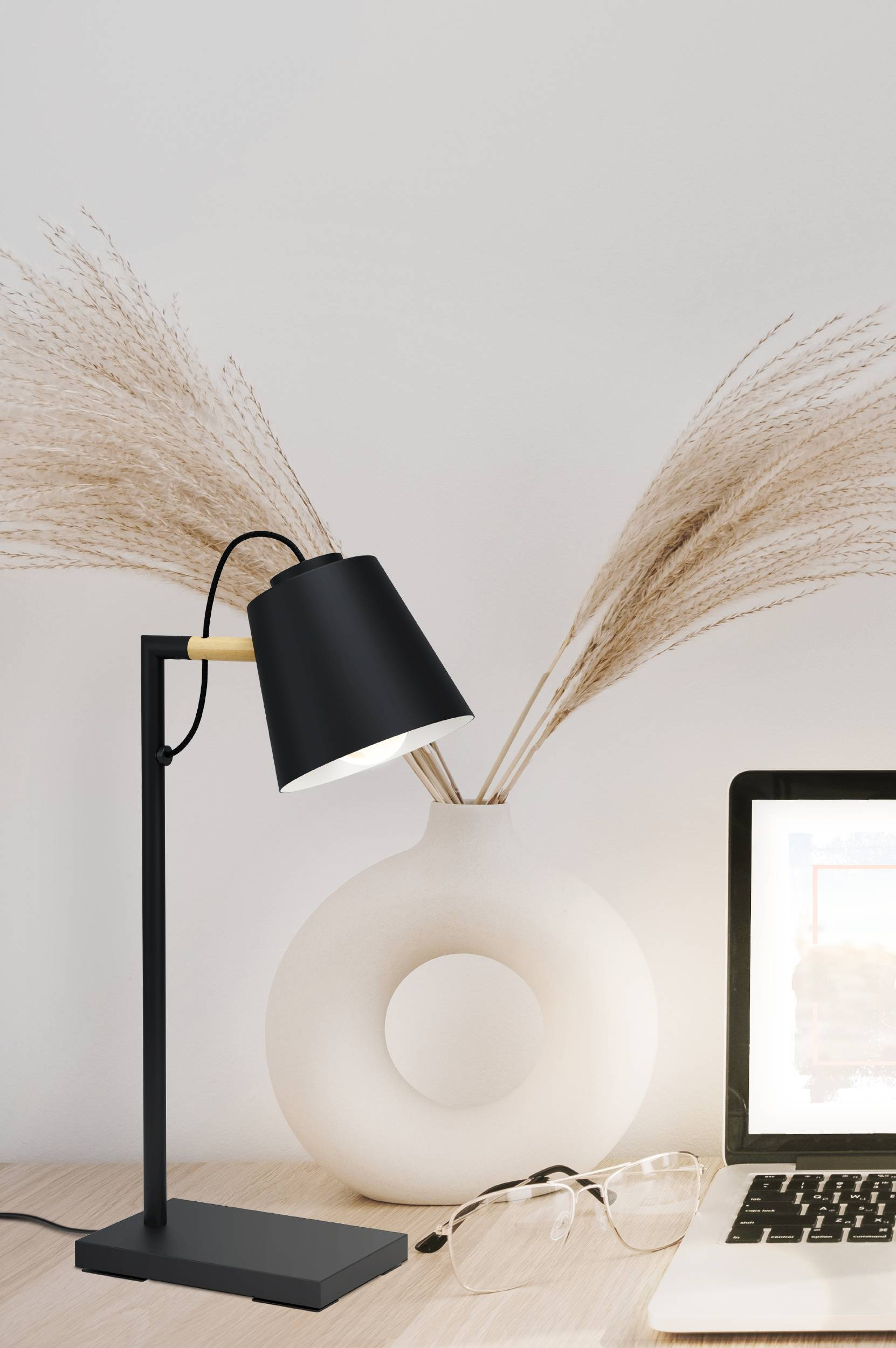 Eine schwarze Schreibtischlampe steht auf einem Schreibtisch mit einem Laptop, einer Vase und einer Brille.