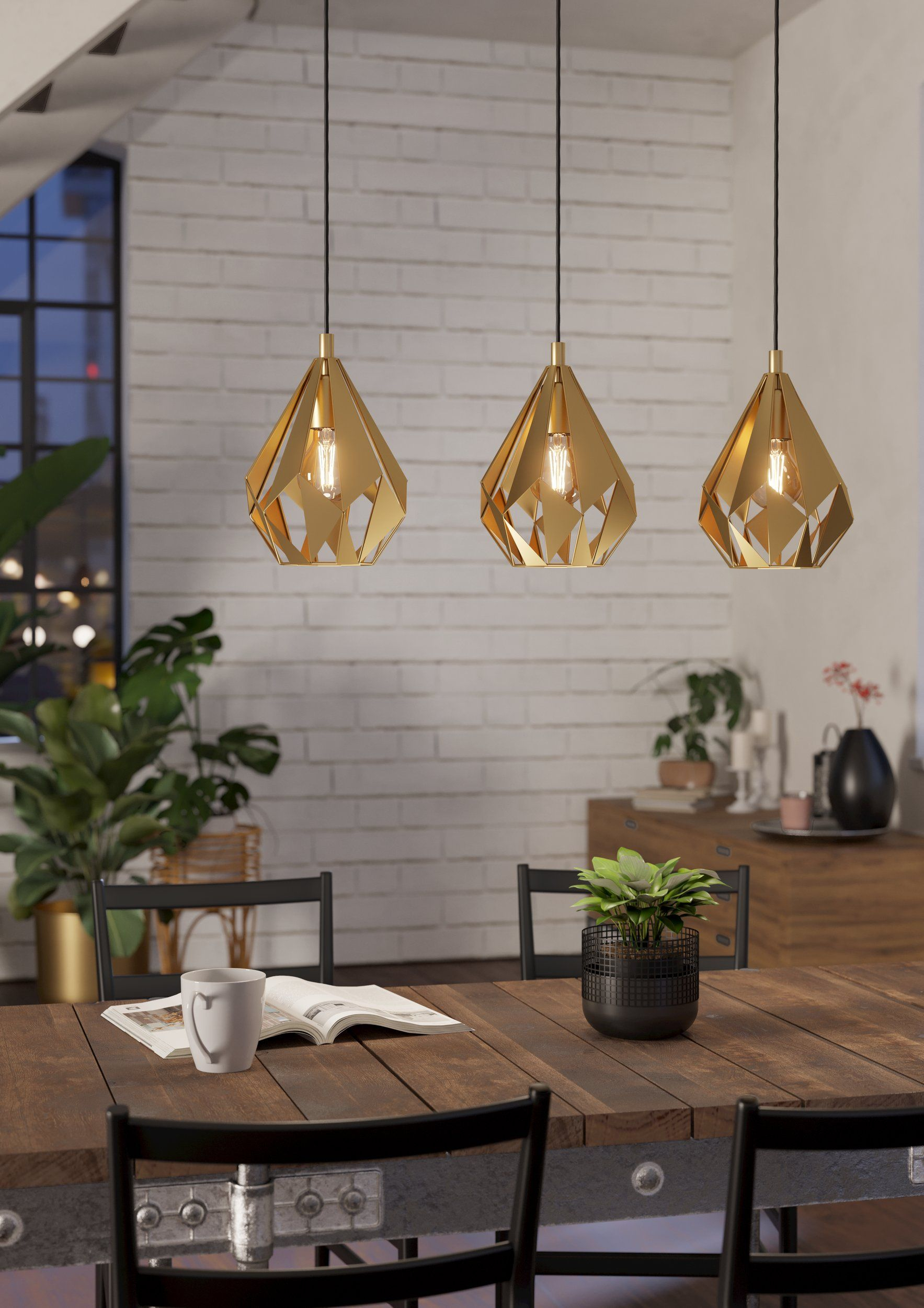 Drei goldene Pendelleuchten mit geometrischen Schirmen, die über einem Holztisch hängen. Hintergrund mit Ziegelmauer.