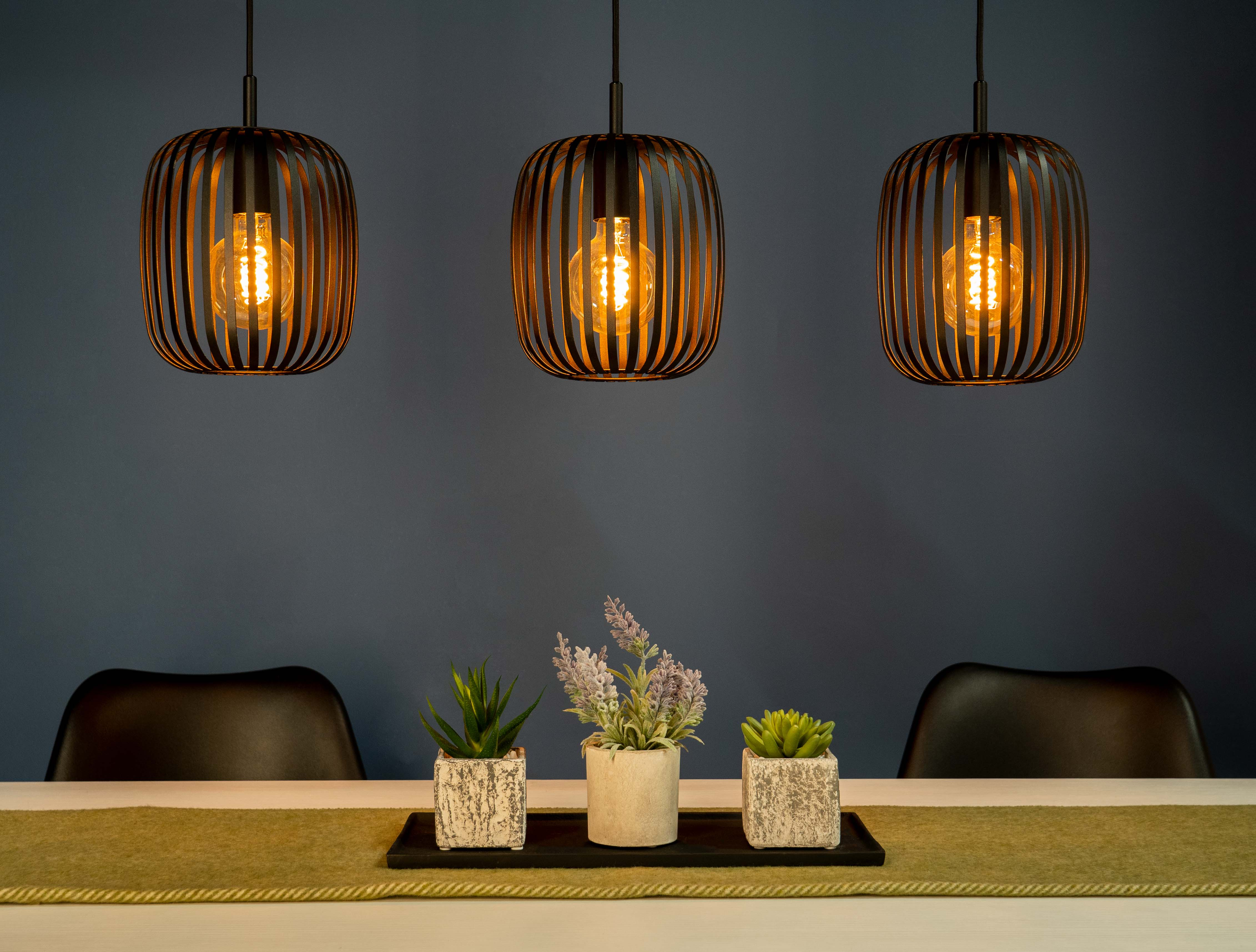 Drie hanglampen met kooikappen hangen boven een tafel. De lichten zijn aan, en er zijn planten en kaarsen.