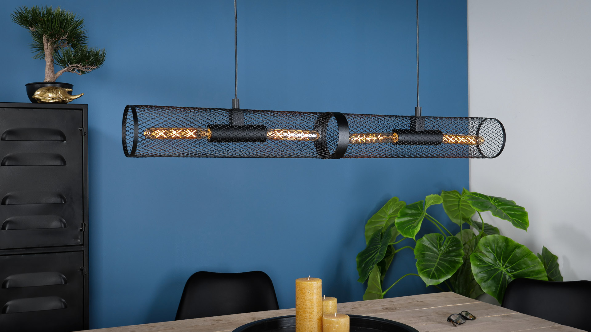 Eine schwarze Metallgitter-Pendelleuchte mit vier Glühbirnen hängt über einem Holztisch.