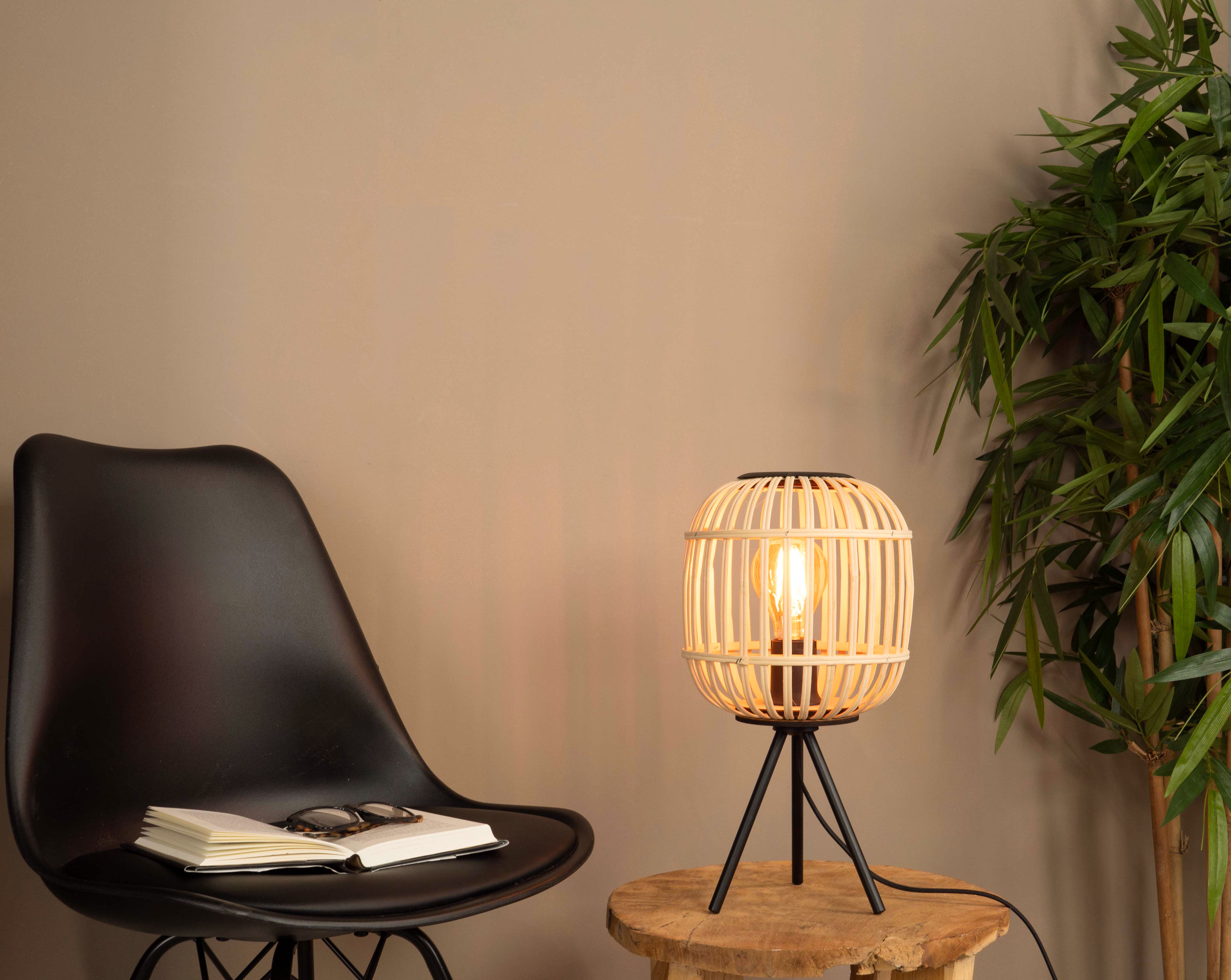 Lampe mit Bambusschatten auf einem Holztisch. Ein schwarzer Stuhl, Buch und Brille im Bild.