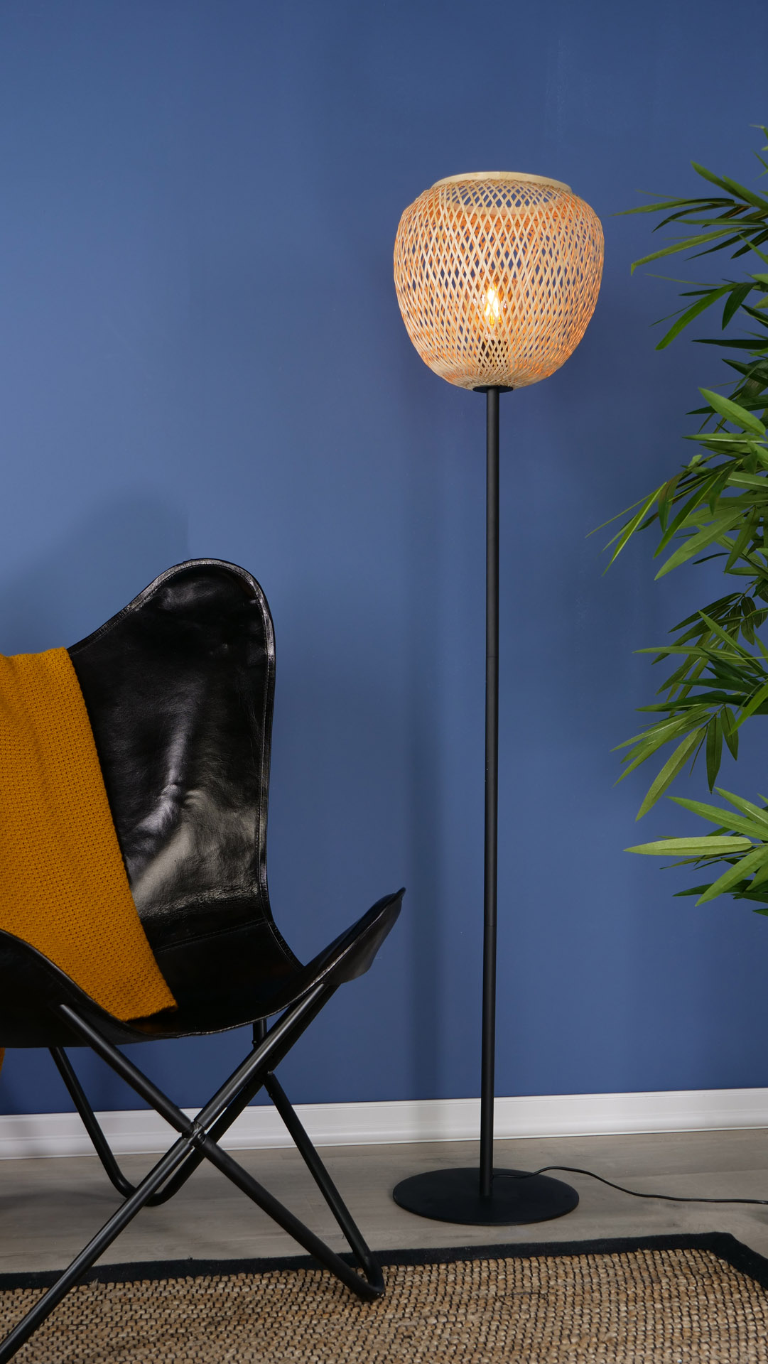 Hohe Stehlampe mit Bambusschirm, neben schwarzem Stuhl und grüner Pflanze an blauer Wand.
