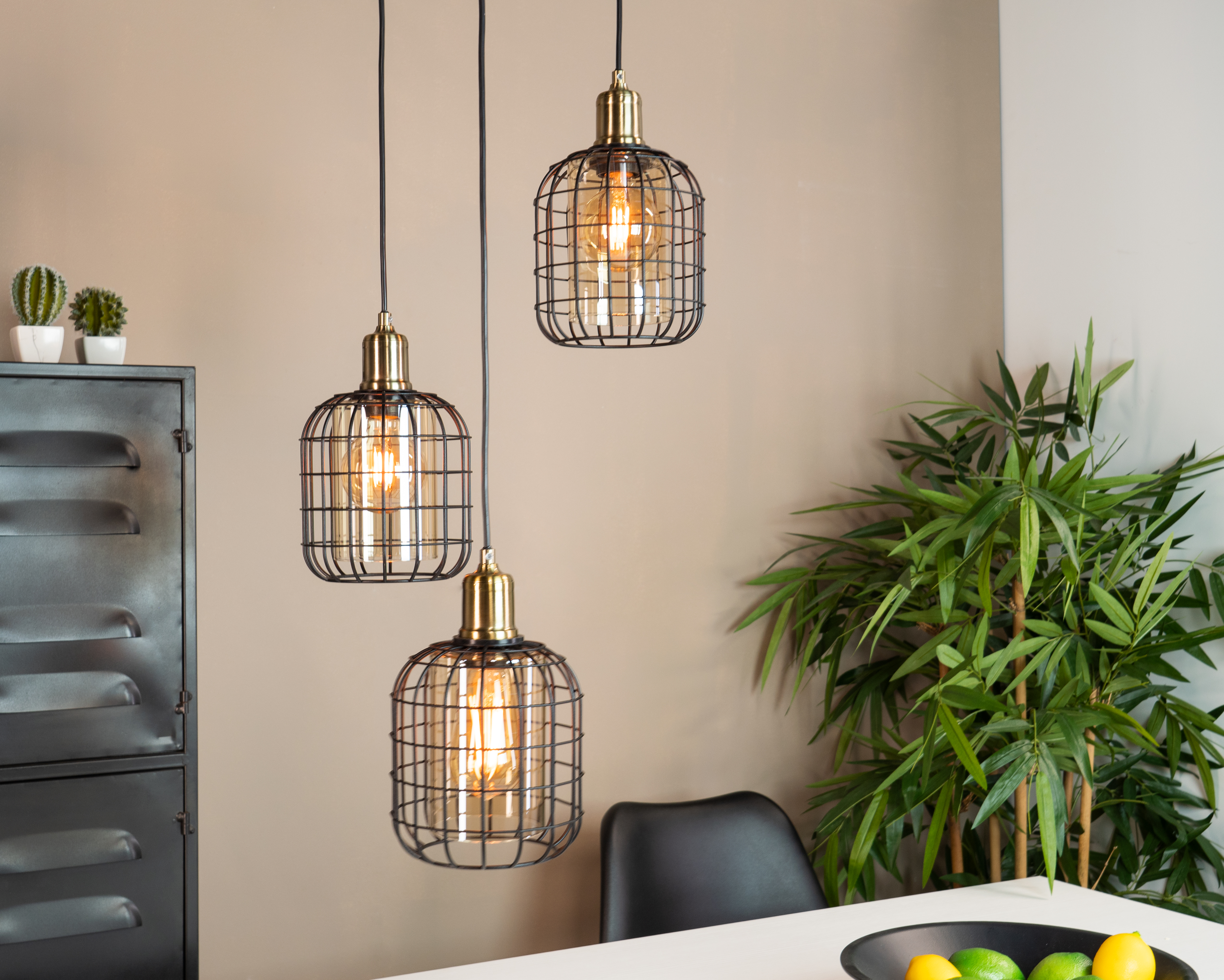 Tre lampade a sospensione sopra un tavolo. Caratterizzate da lampadine Edison in gabbia nera. Ambiente stanza.