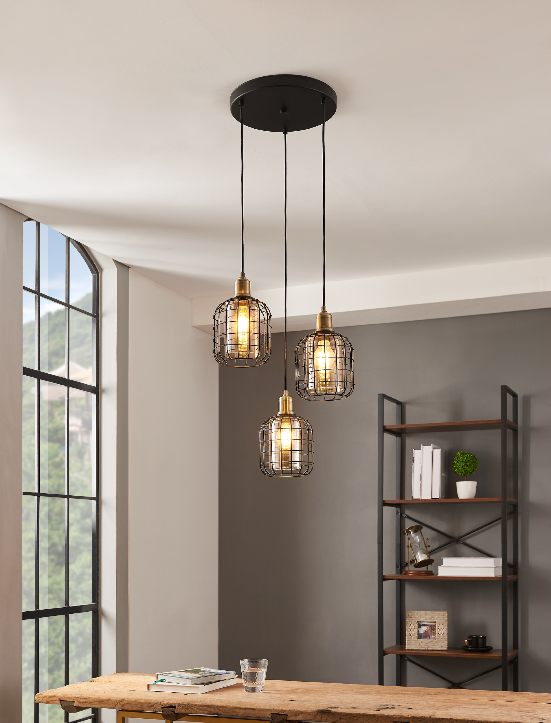 Tre lampade a sospensione con lampadine Edison sopra un tavolo. Lo sfondo include una finestra e una mensola.