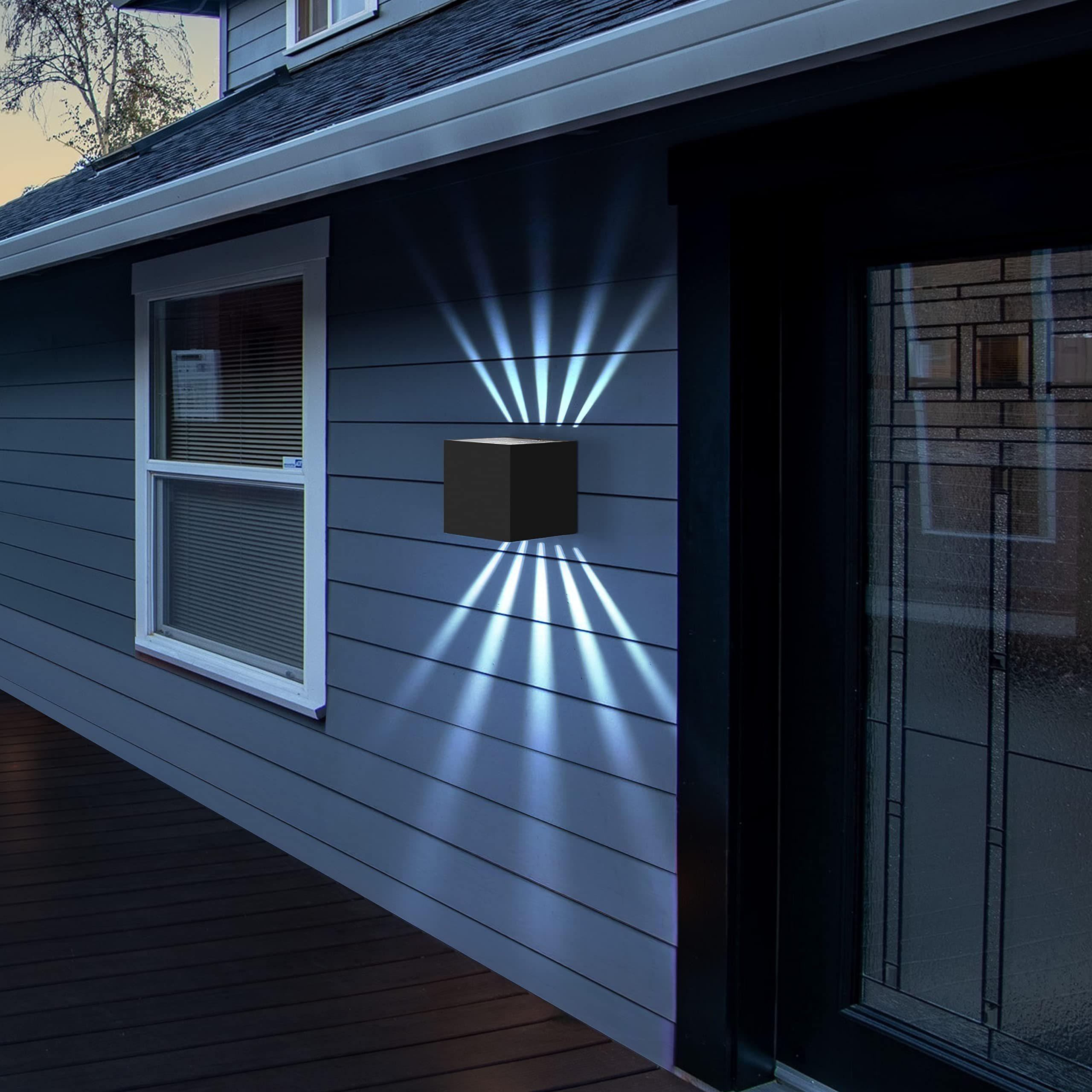 Una lampada da parete nera a forma di cubo è montata sull'esterno di una casa blu, emettendo fasci di luce.