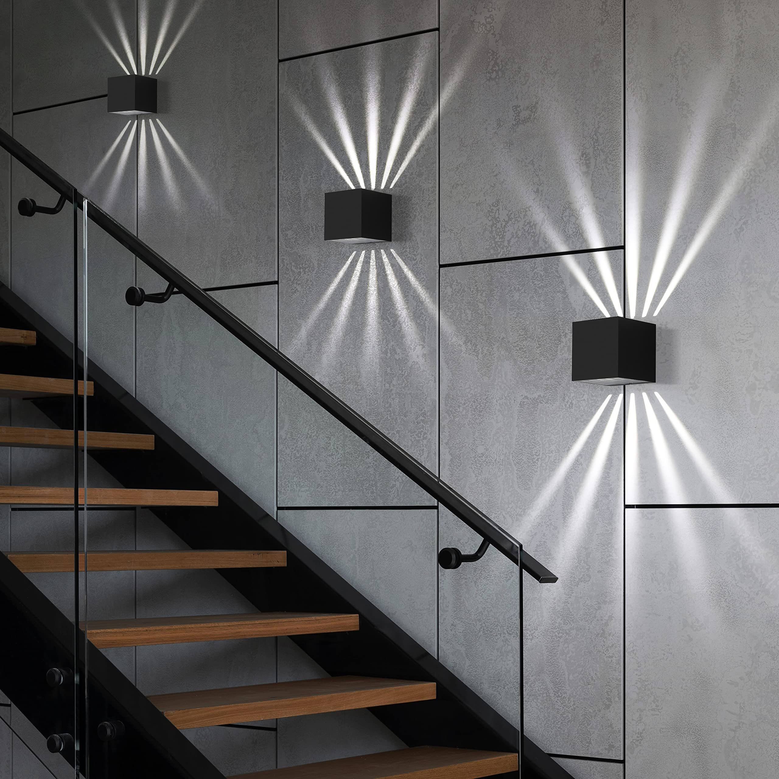 Tre lampade da parete nere a forma di cubo emettono fasci di luce su un vano scale con scale in legno e una ringhiera in vetro.