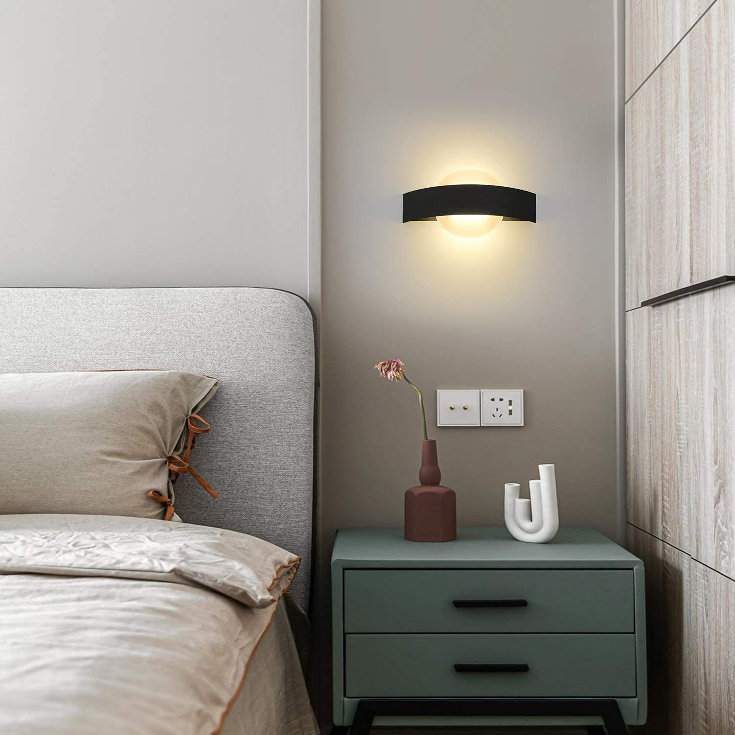 Una scena di camera da letto con una lampada da parete, un letto, un comodino con un vaso e decorazioni e un armadio in legno.