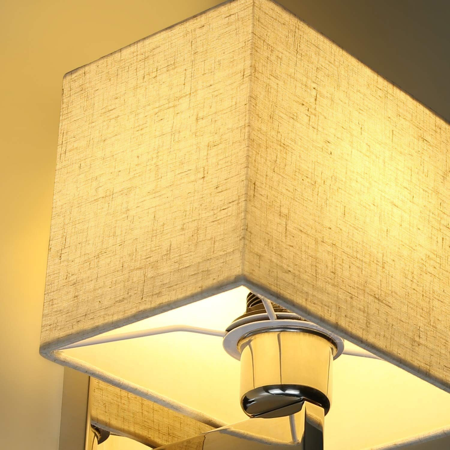 Viene mostrato un primo piano di una lampada da parete illuminata con paralume quadrato in tessuto e base argentata.