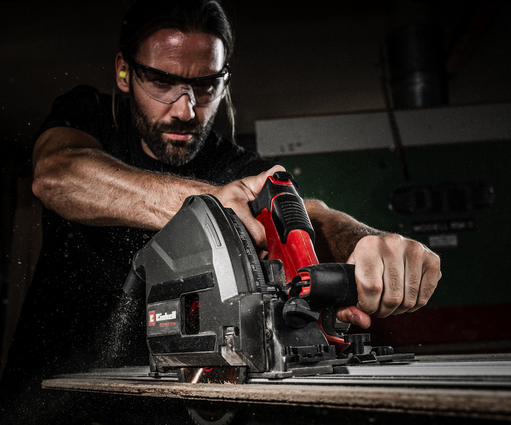 Ein Mann mit Schutzbrille benutzt eine rot-graue Einhell-Tauchsäge, um in einer Werkstatt ein Holzbrett zu sägen.