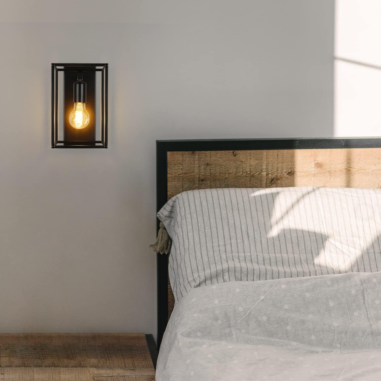 Czarna lampa ścienna zamontowana na białej ścianie. W tle łóżko z drewnianym zagłówkiem.