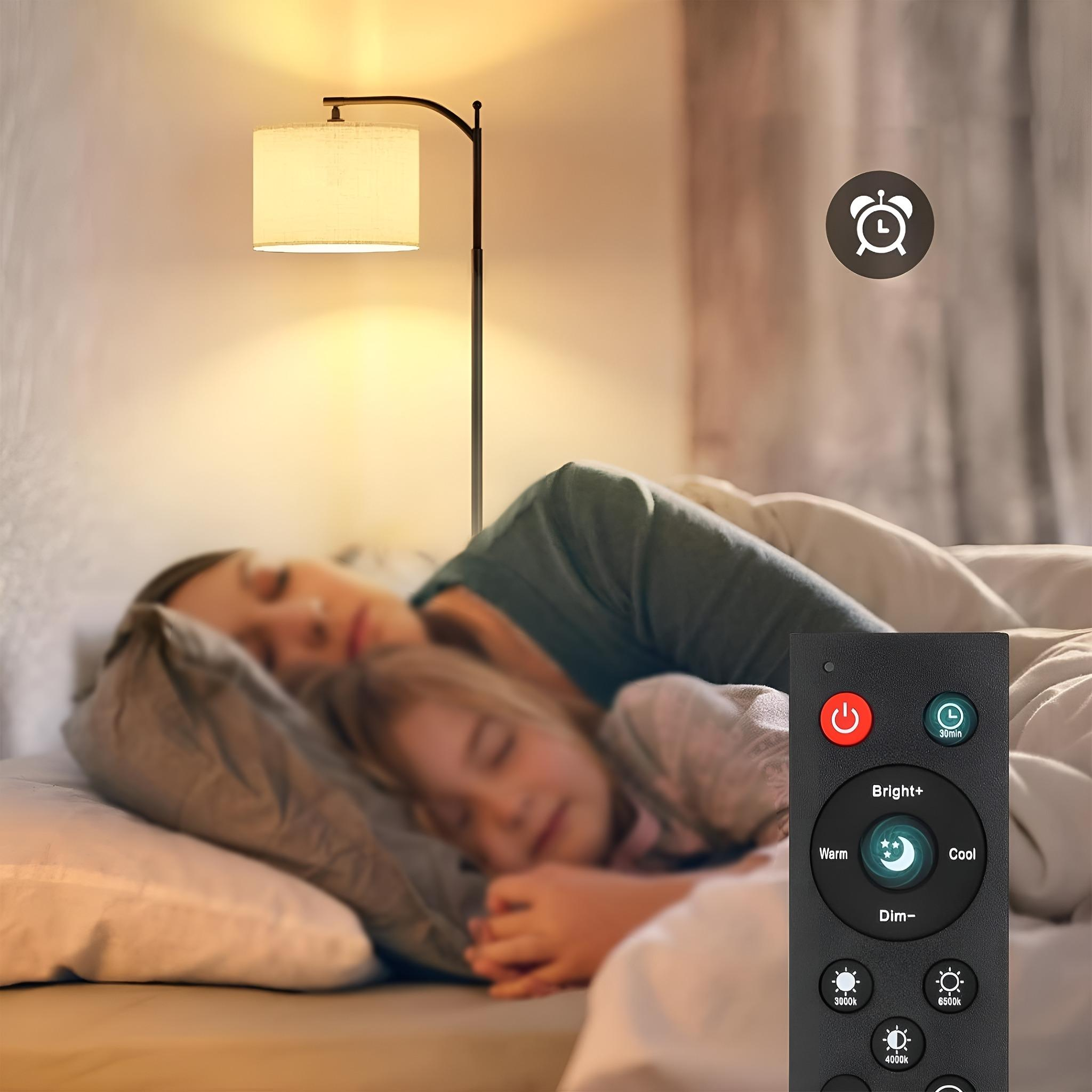 Eine Stehlampe mit Lampenschirm neben einem Bett. Zwei Personen schlafen. Fernbedienung im Vordergrund.