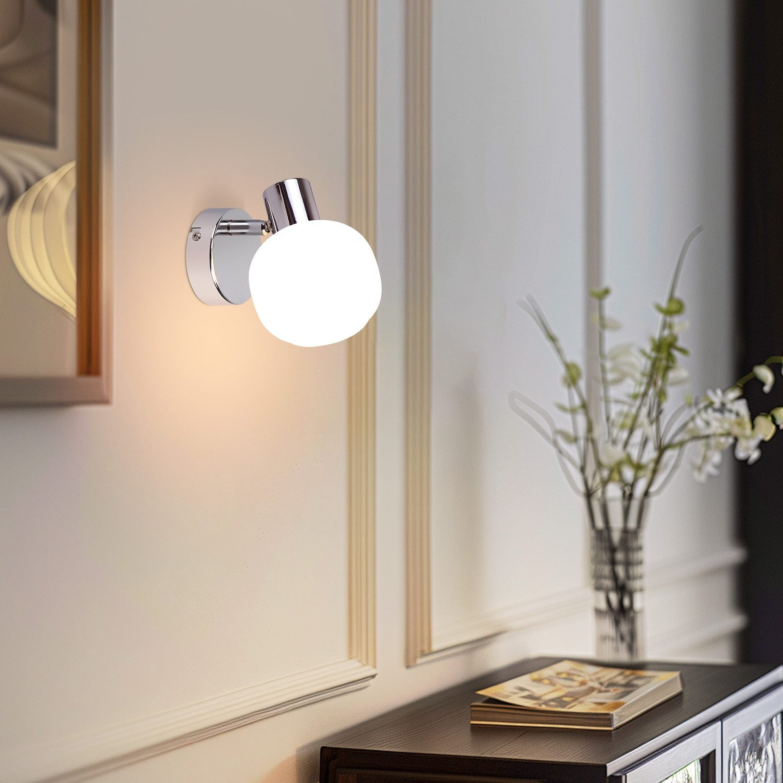 Una lampada da parete cromata con paralume a globo bianco illumina una parete con un'immagine incorniciata e un vaso di fiori.