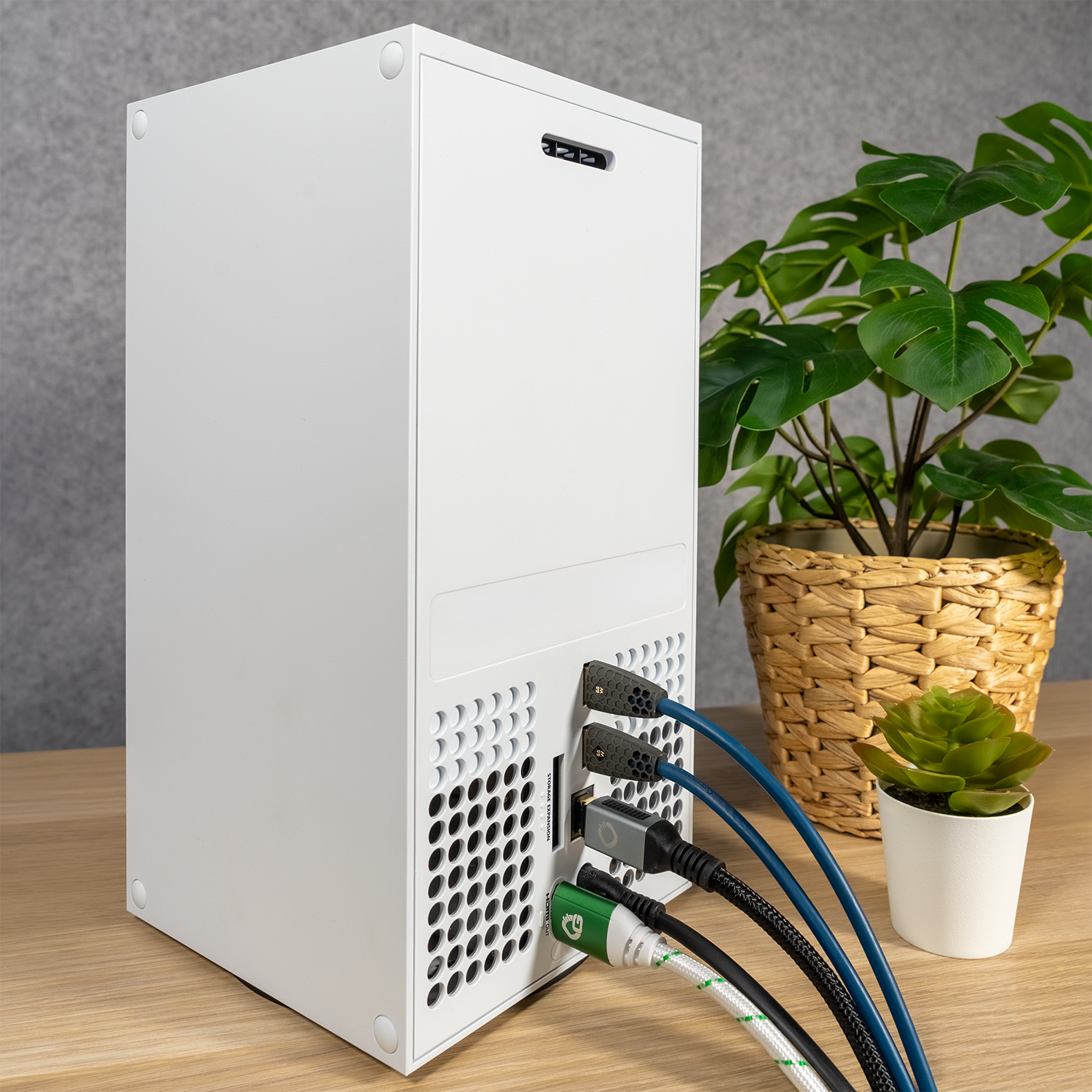 Eine Spielekonsole mit angeschlossenen Kabeln. Ein grün-weißes HDMI-Kabel ist sichtbar. Eine Pflanze ist im Hintergrund.