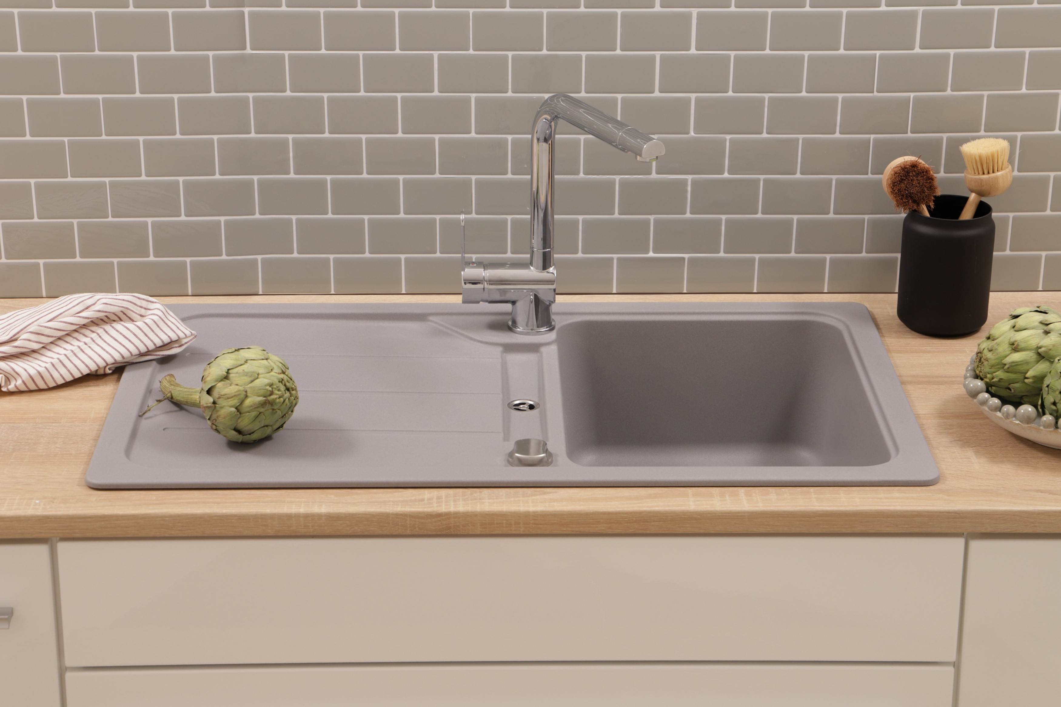 Ein Küchenbecken mit Wasserhahn, Artischocke und Geschirrtuch. Der Fliesenspiegel ist grauer Kacheln, die Arbeitsplatte aus Holz.
