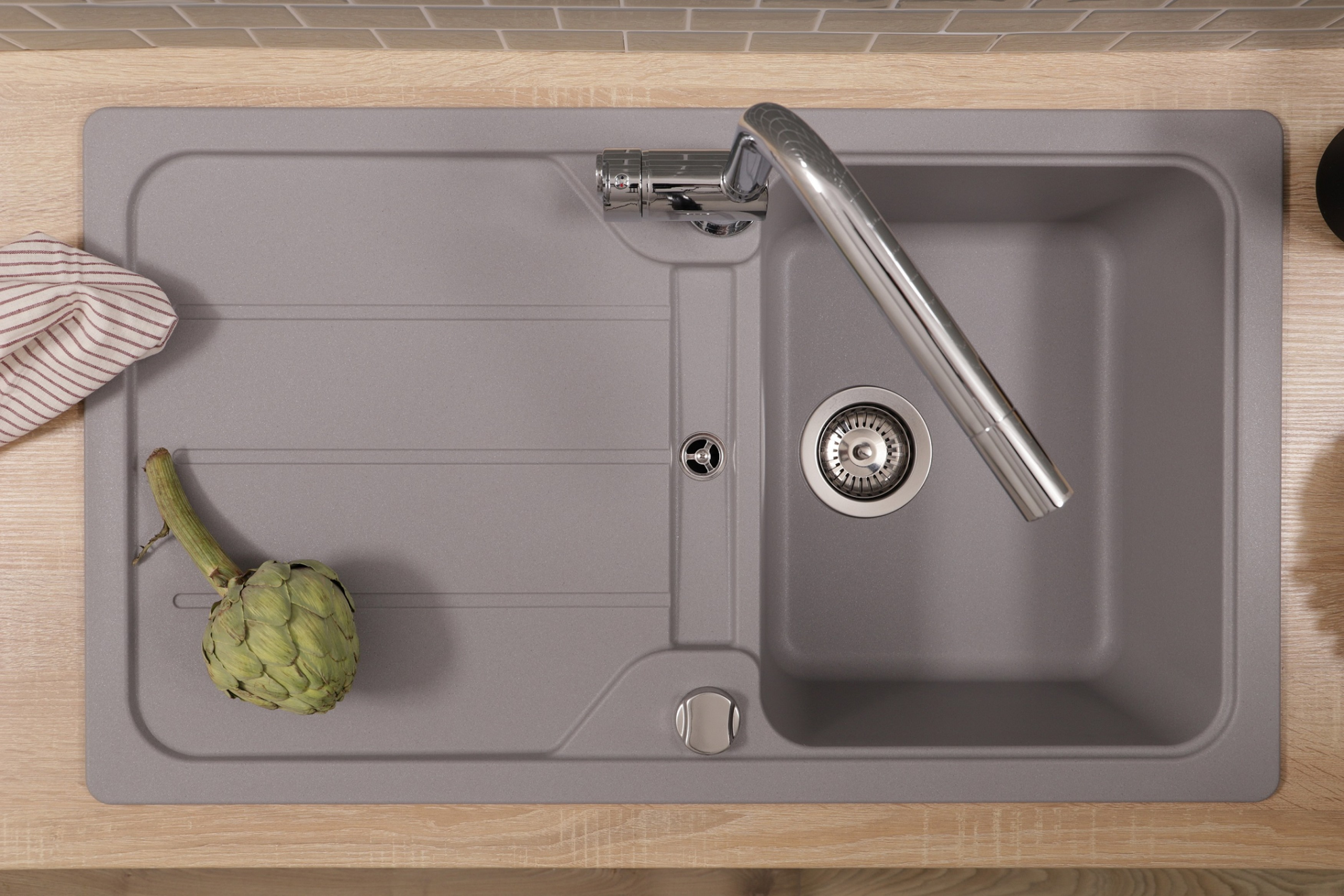Ein Küchenbecken mit Wasserhahn, Artischocke und Geschirrtuch. Der Fliesenspiegel ist grauer Kacheln, die Arbeitsplatte aus Holz.
