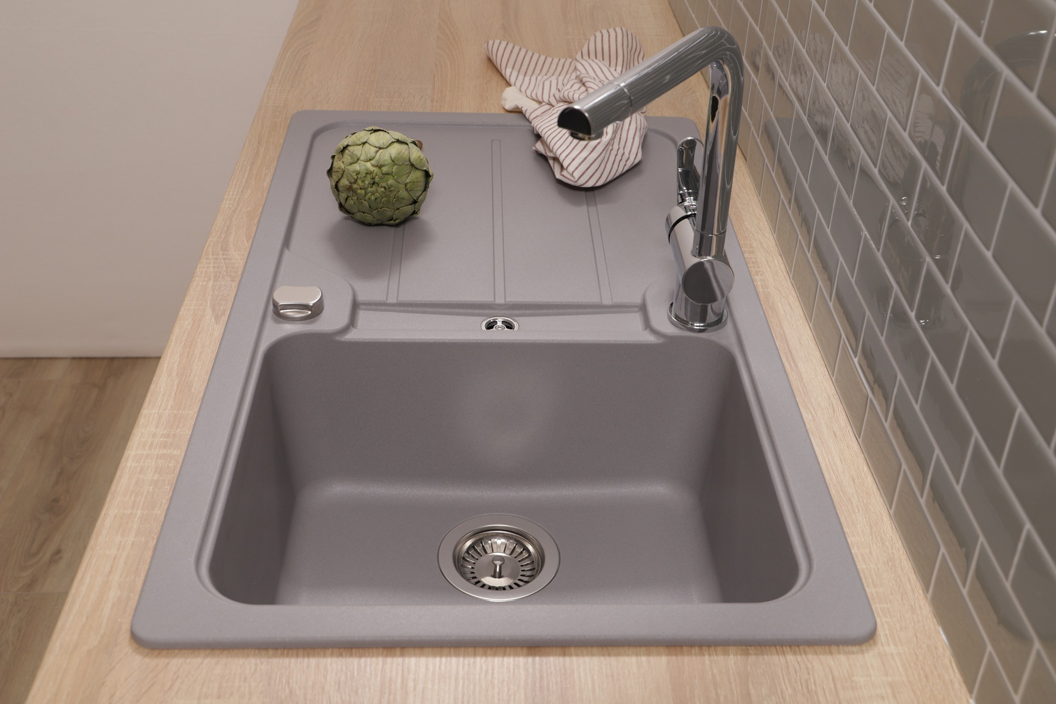 Ein Küchenbecken mit Wasserhahn, Artischocke und Geschirrtuch. Der Fliesenspiegel ist grauer Kacheln, die Arbeitsplatte aus Holz.