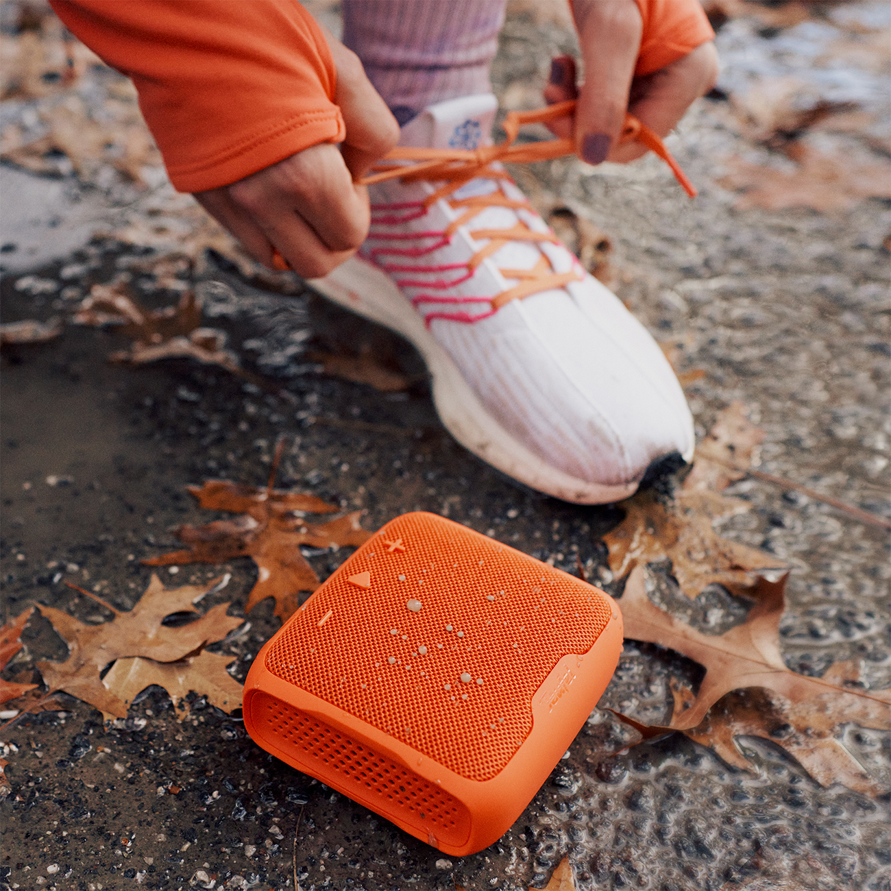 Persoon die een veter vastmaakt aan een wit-oranje hardloopschoen bij een oranje luidspreker.