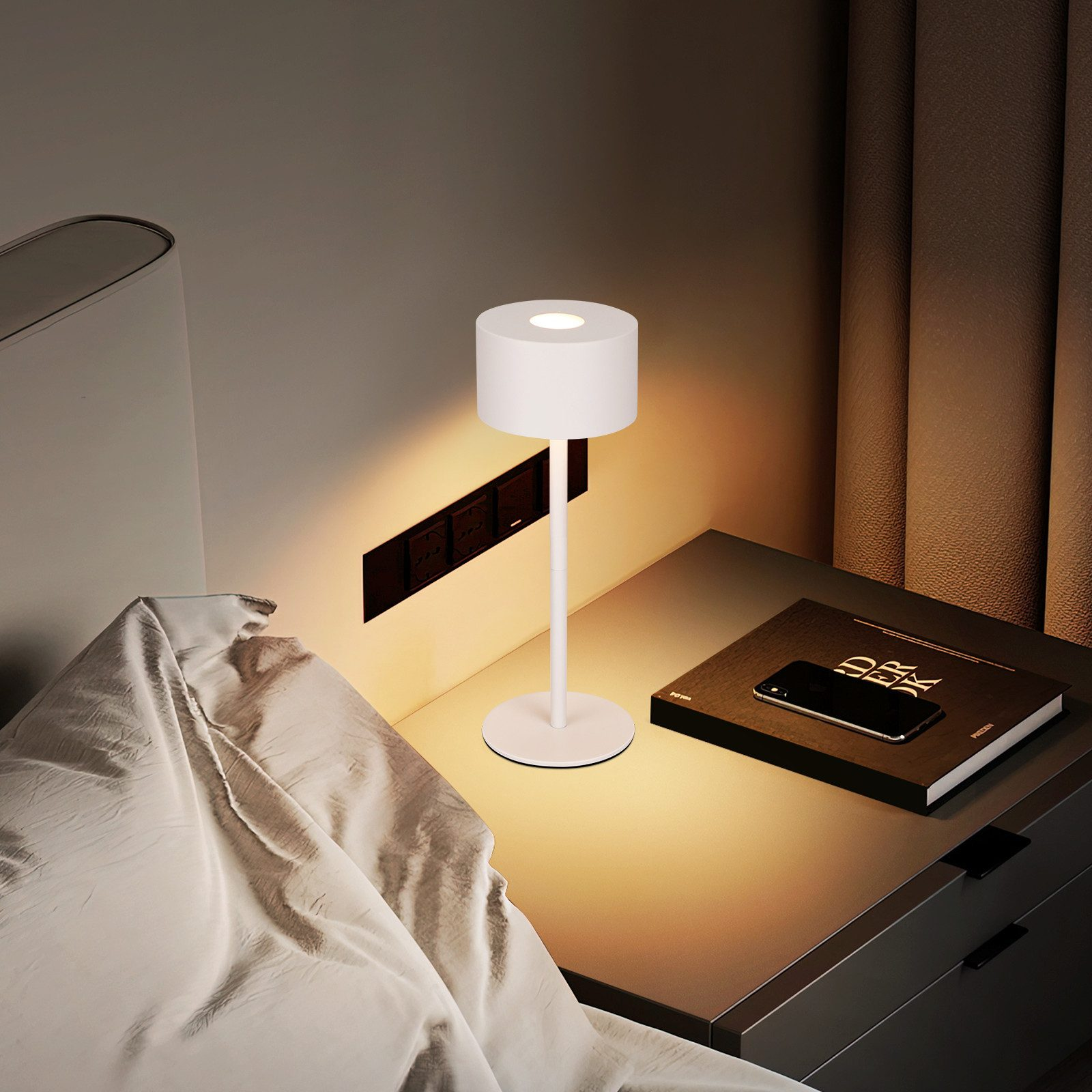 Biała lampa na szafce nocnej obok łóżka. Na stole leżą również książka i telefon.