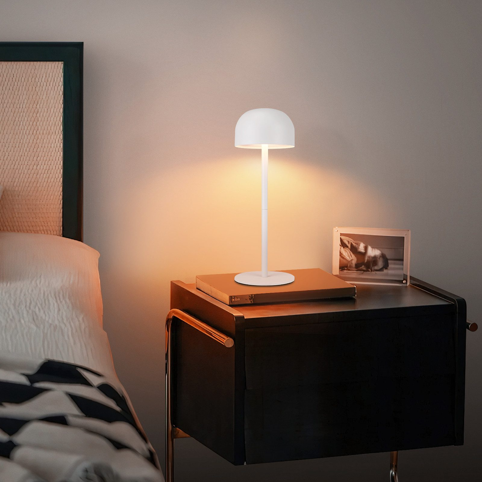 Biała lampa stoi na czarnym stoliku nocnym. Na stoliku nocnym znajduje się oprawione zdjęcie i książka.