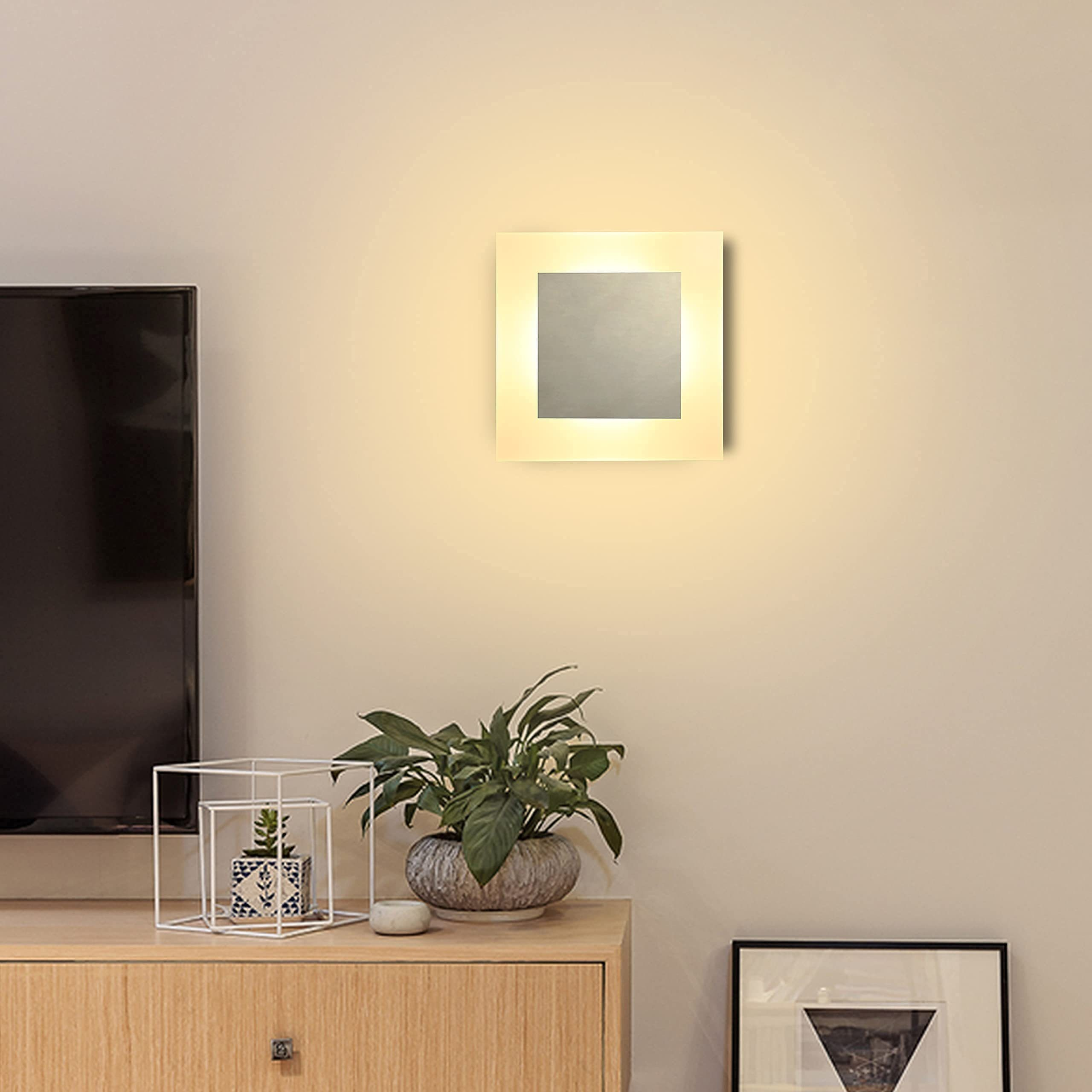 Una luce da parete quadrata con un centro argentato, montata sopra un mobile in legno con piante.