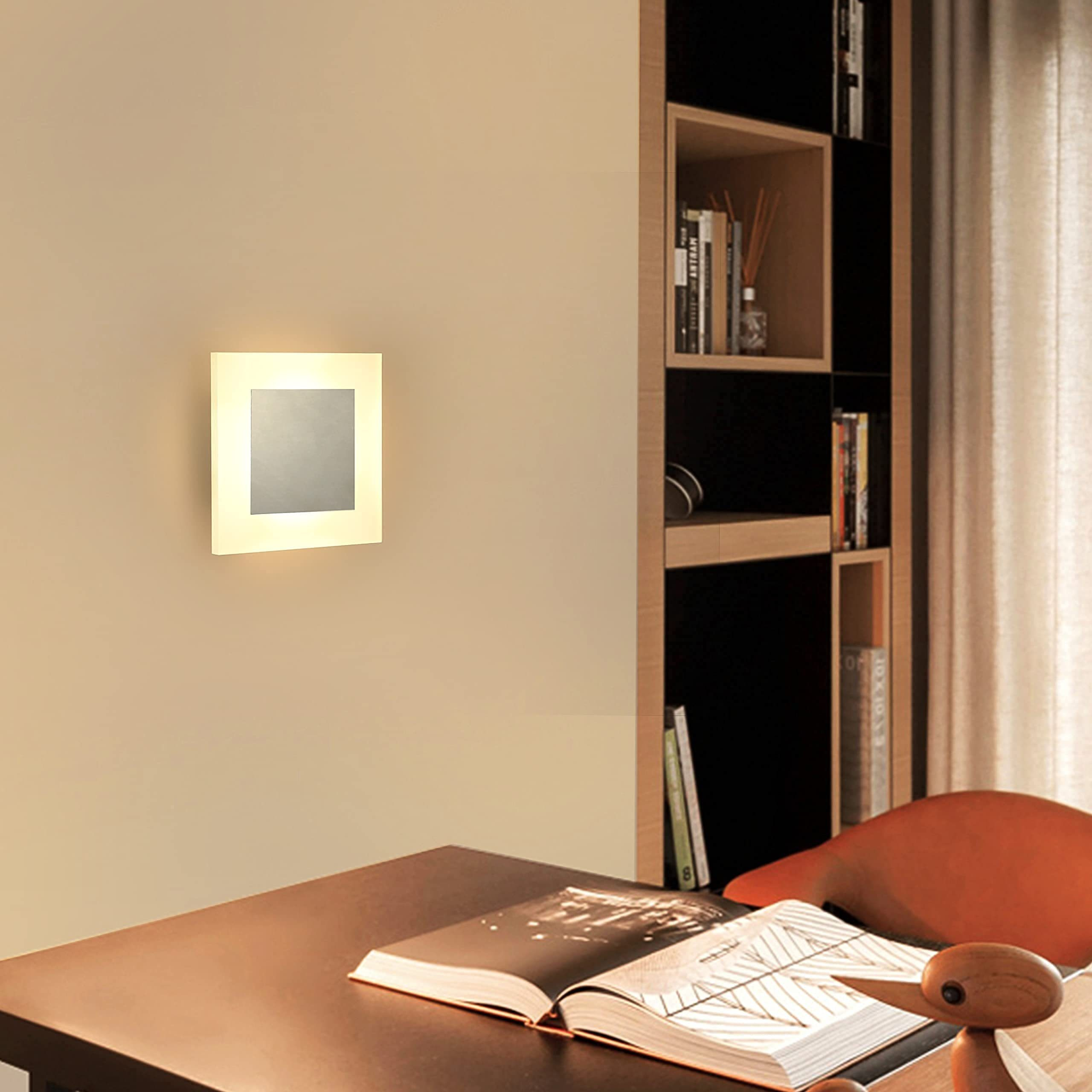 Una lampada da parete quadrata illuminata illumina una stanza con un libro su una scrivania e una mensola sullo sfondo.
