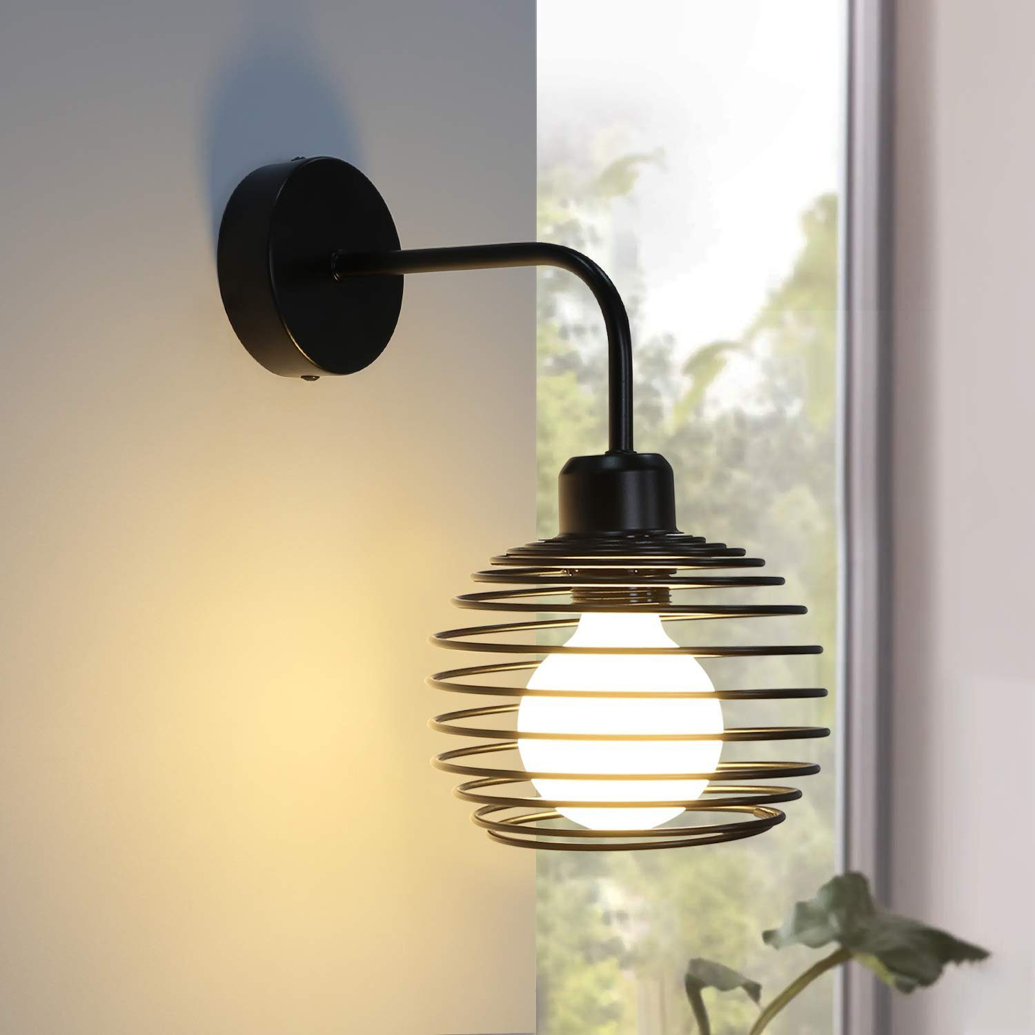 Lampada da parete nera a gabbia con lampadina accesa, vicino a una finestra e una pianta.