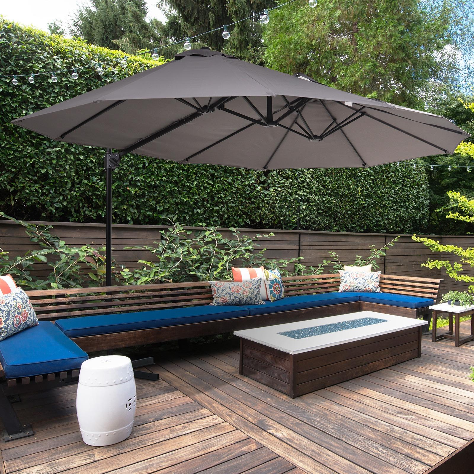 Ein grauer Doppelsonnenschirm beschattet eine Terrasse mit Sitzgelegenheiten, Kissen, einer Feuerstelle und üppigem Grün im Hintergrund.