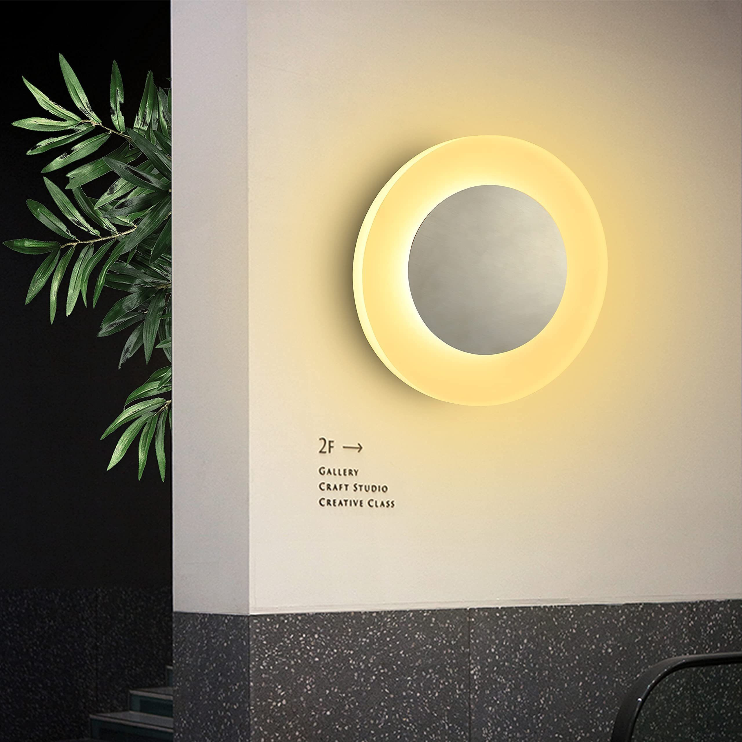 Interno con una lampada da parete circolare accanto a una pianta, con indicazioni per una galleria e uno studio.